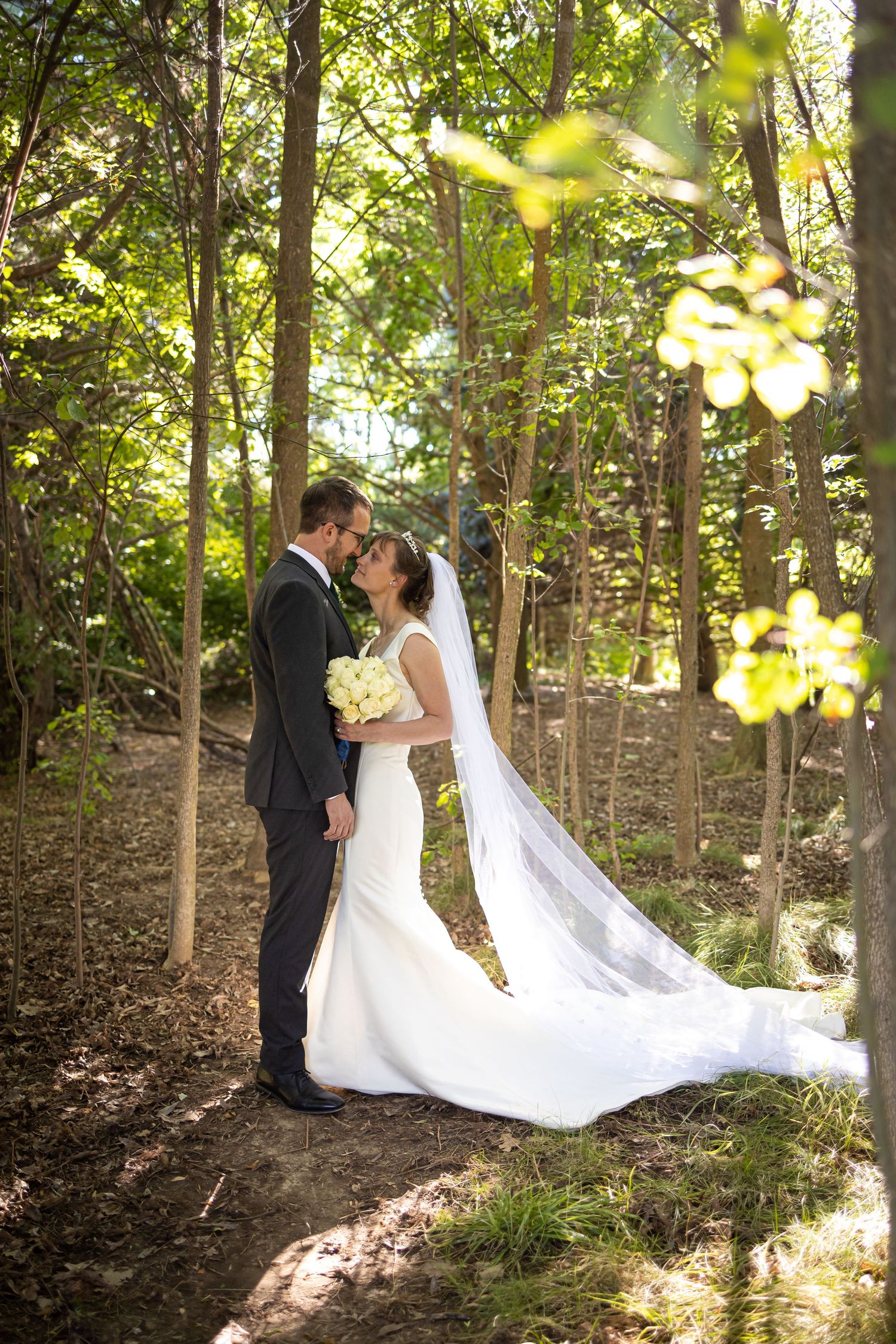 Newlyweds share romantic moment on wooded path with sunlight filtering through trees.