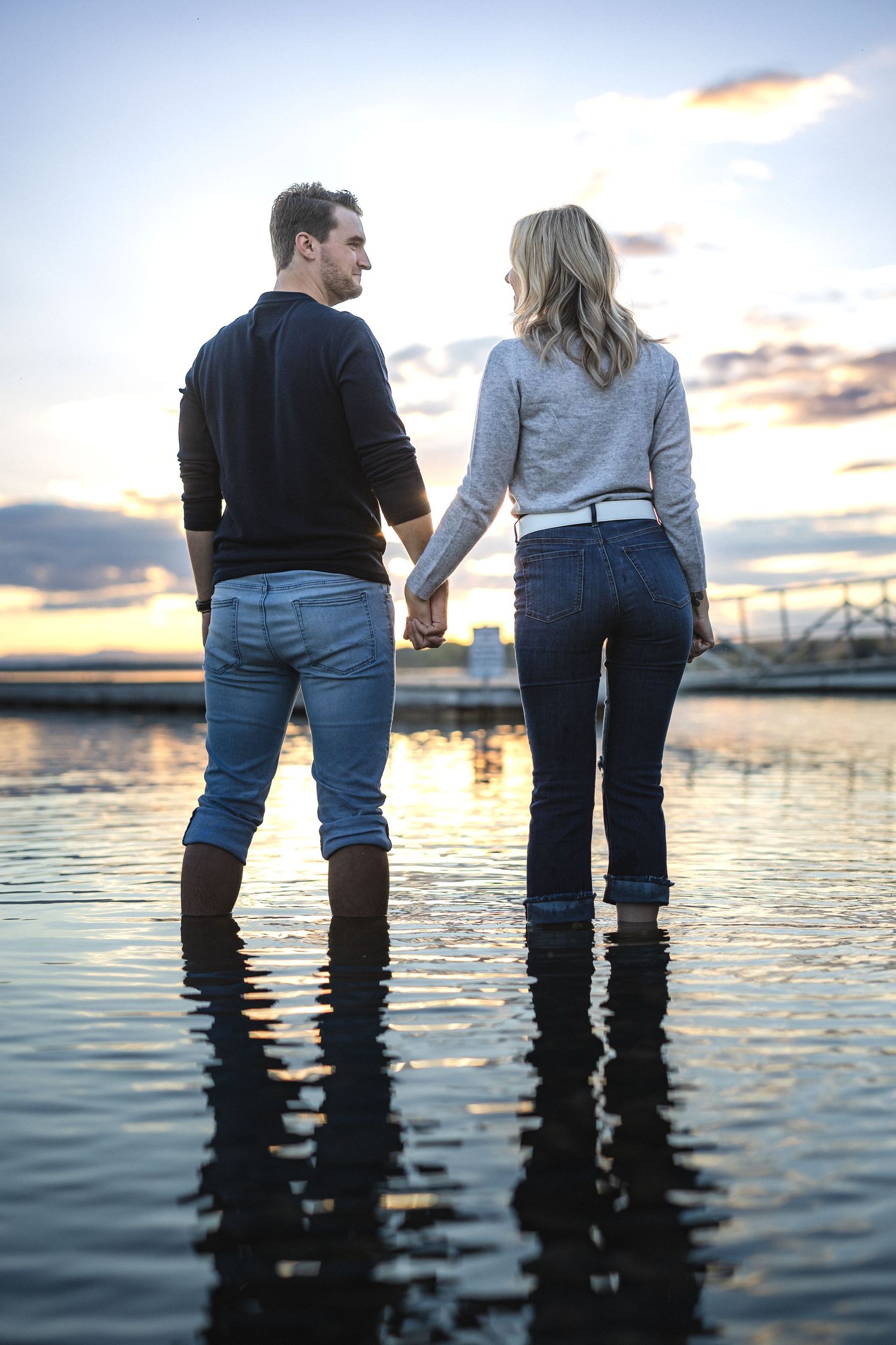 Couple in casual wear holding hands while standing knee-deep in water at dusk.