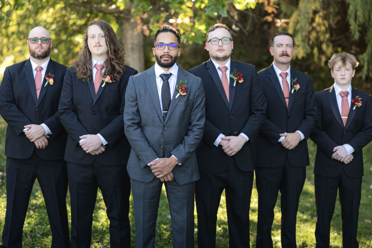 Groomsmen in dark suits with red accents pose outdoors in a formal group photo.