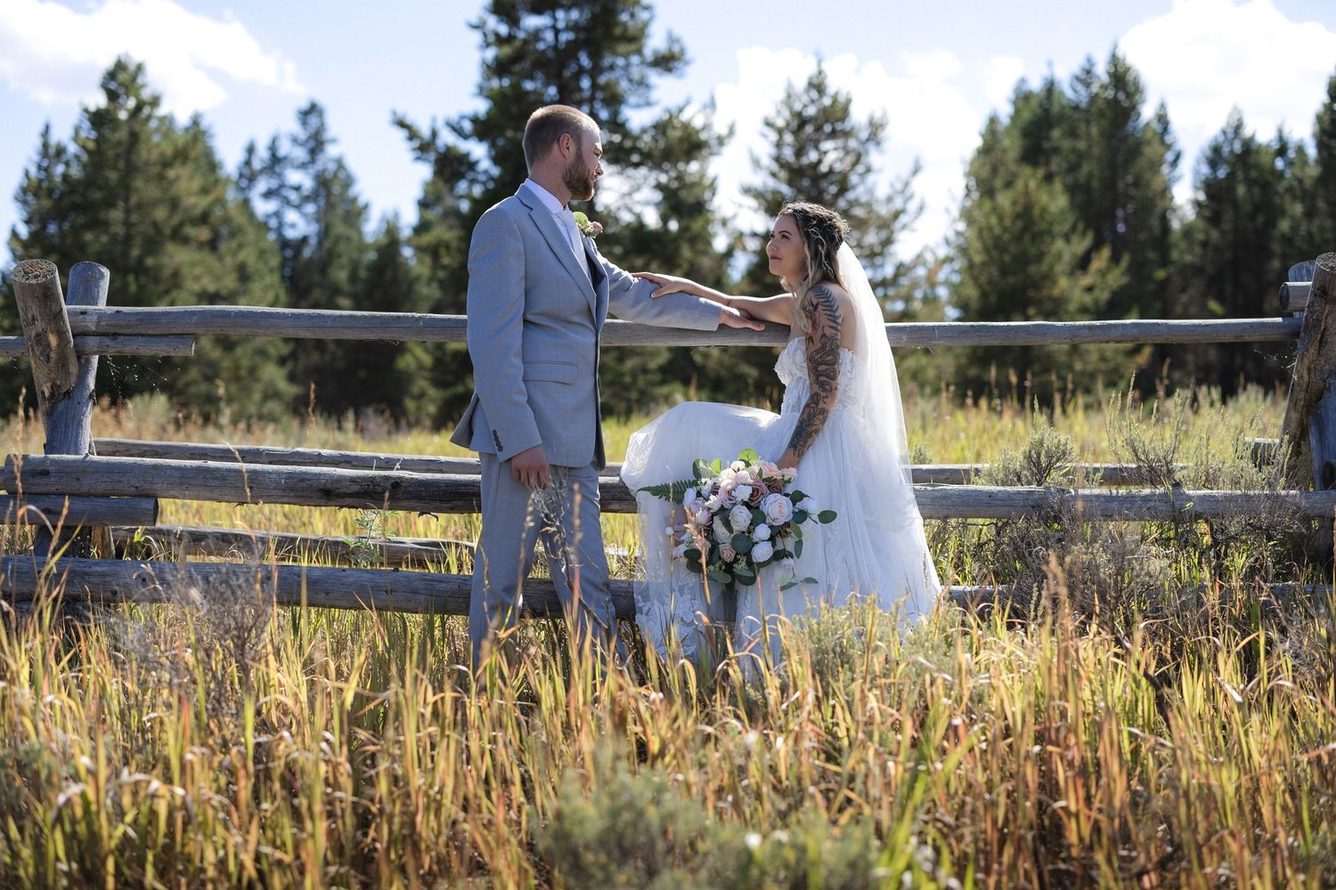 A couple poses along a rustic wooden fence in an outdoor mountain setting.