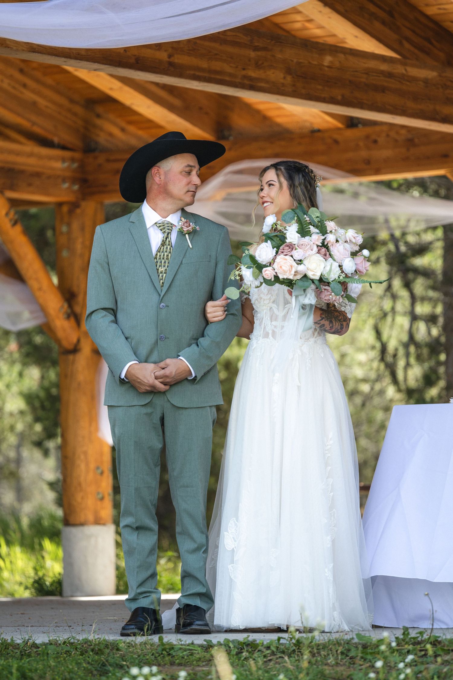 Couple in wedding attire standing under wooden pavilion during rustic outdoor ceremony.
