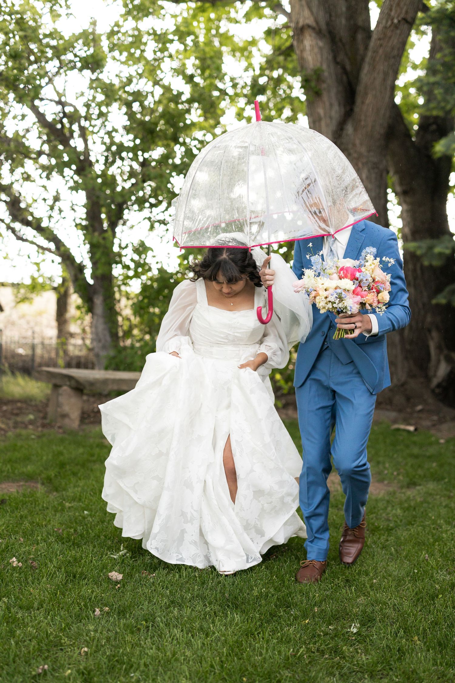 Newlyweds share a playful moment under a clear umbrella in a garden setting with bride in white dress and groom in blue suit.