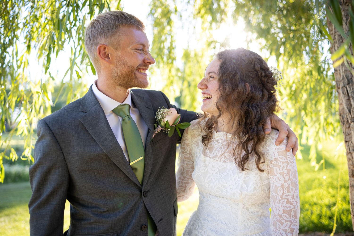 Happy couple share a joyful moment in sunlit garden with willow trees in background.