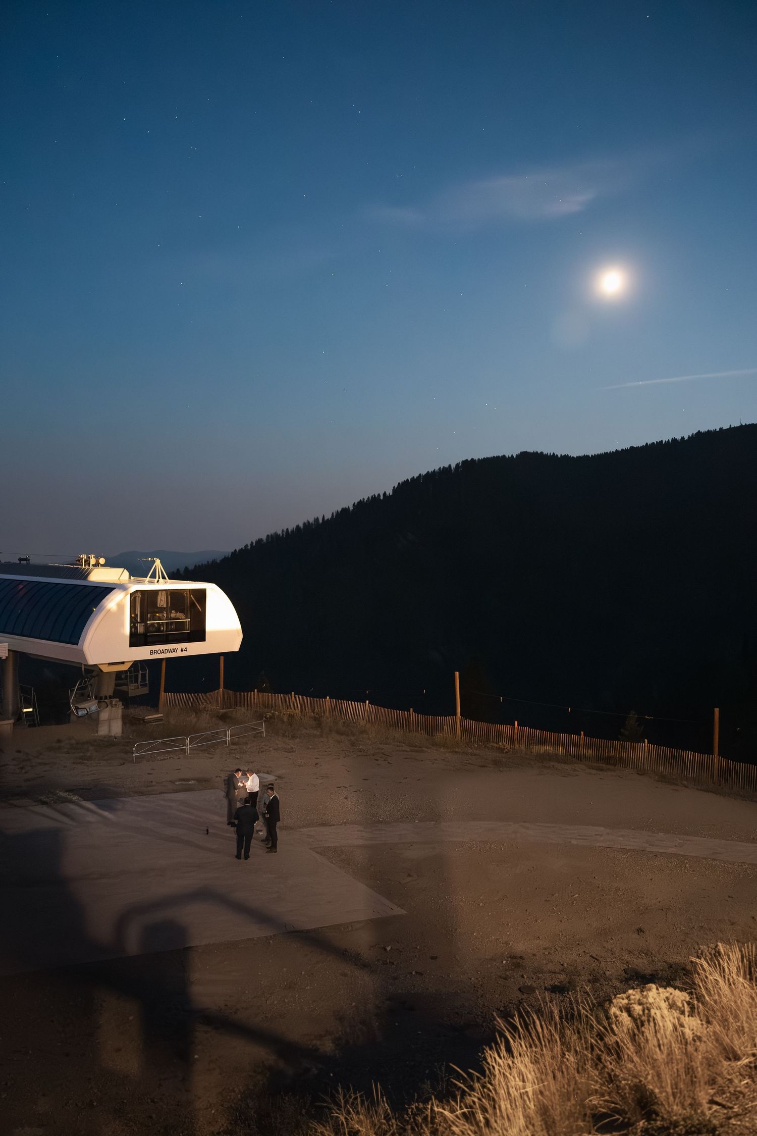 Mountain landscape at dusk with illuminated venue structures and full moon in the background.