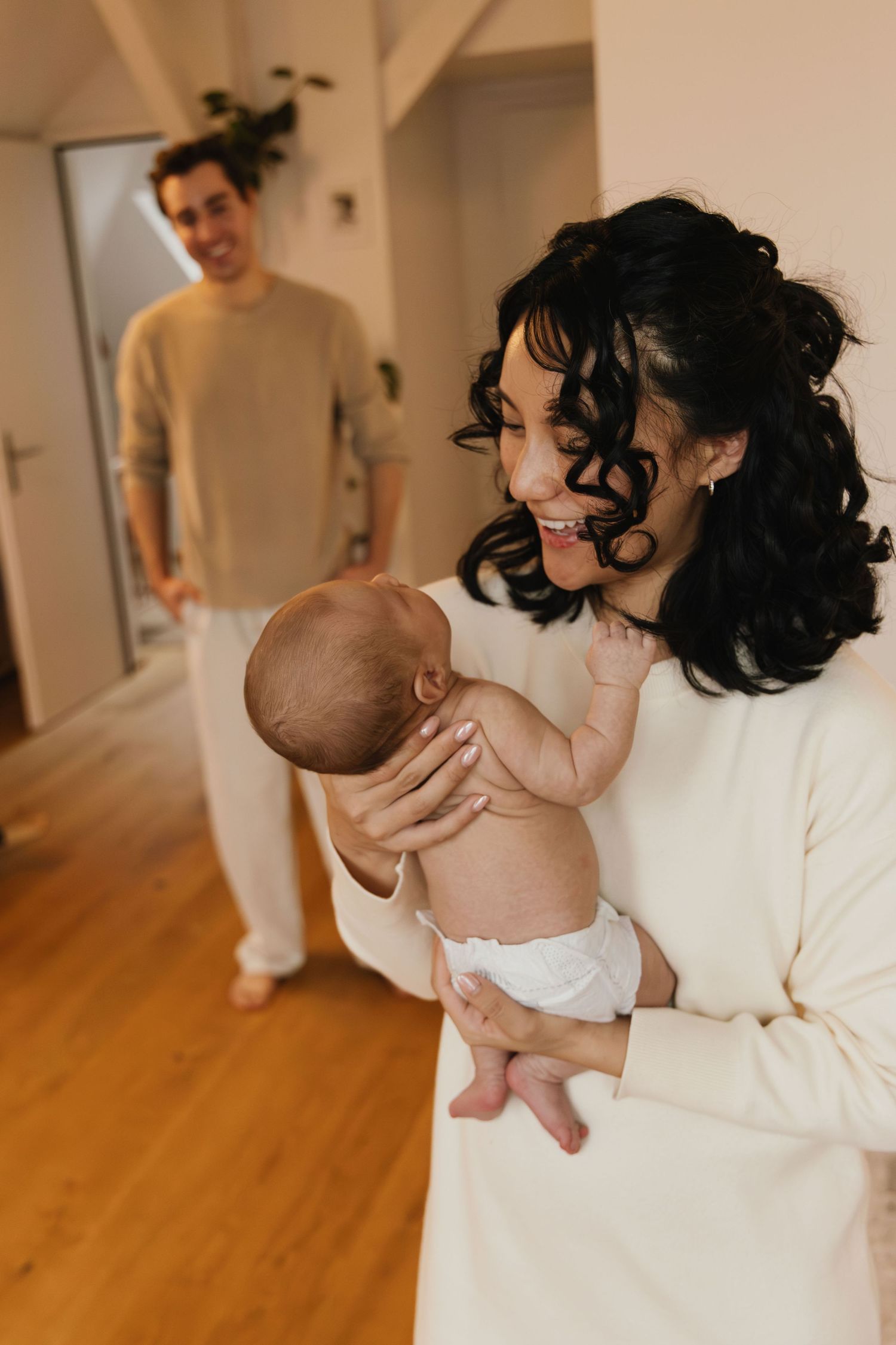 Mother holding her newborn while the father smiles in the background during a family photoshoot.