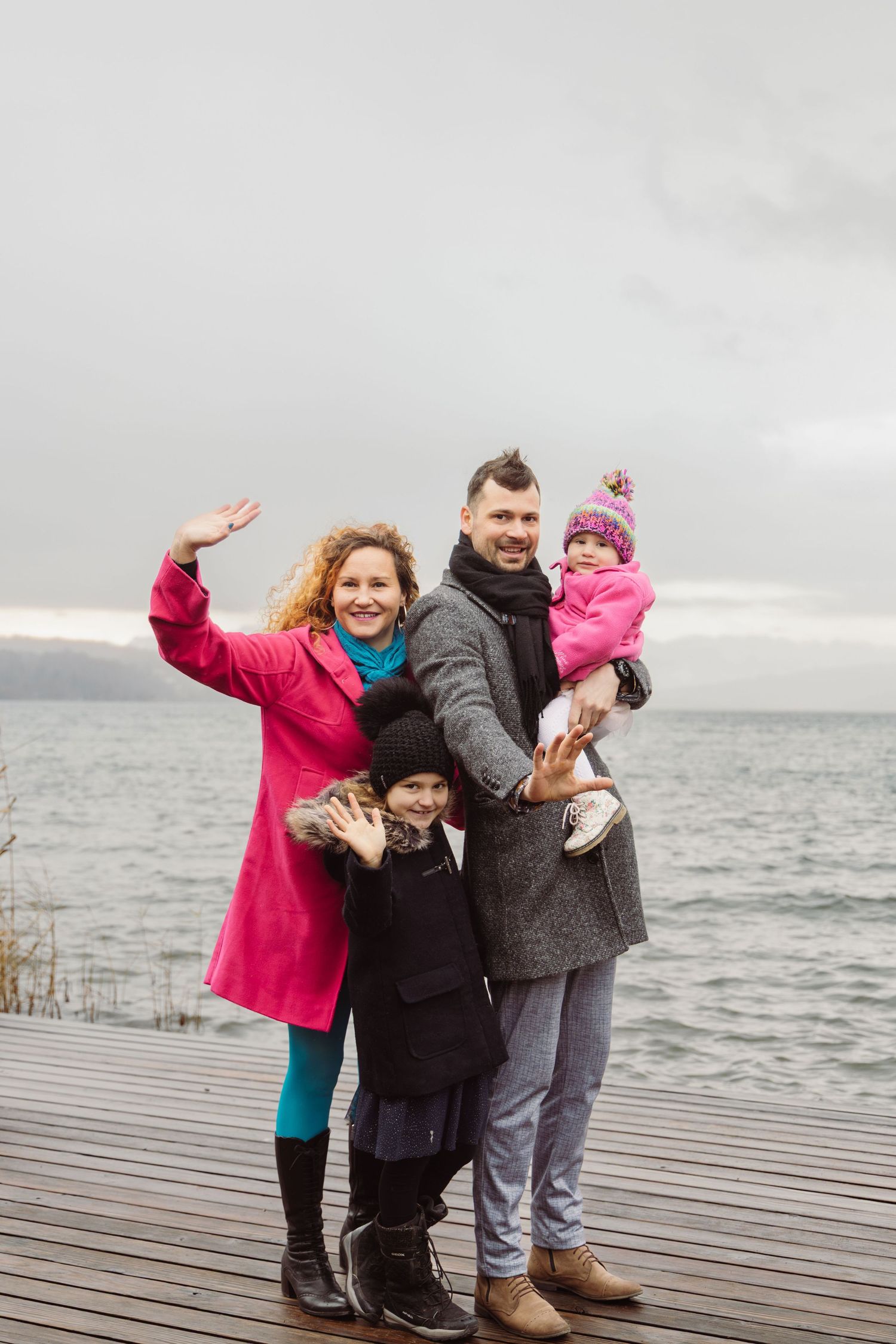 Family waving at the camera with a lake in the background during a winter photoshoot in Switzerland.