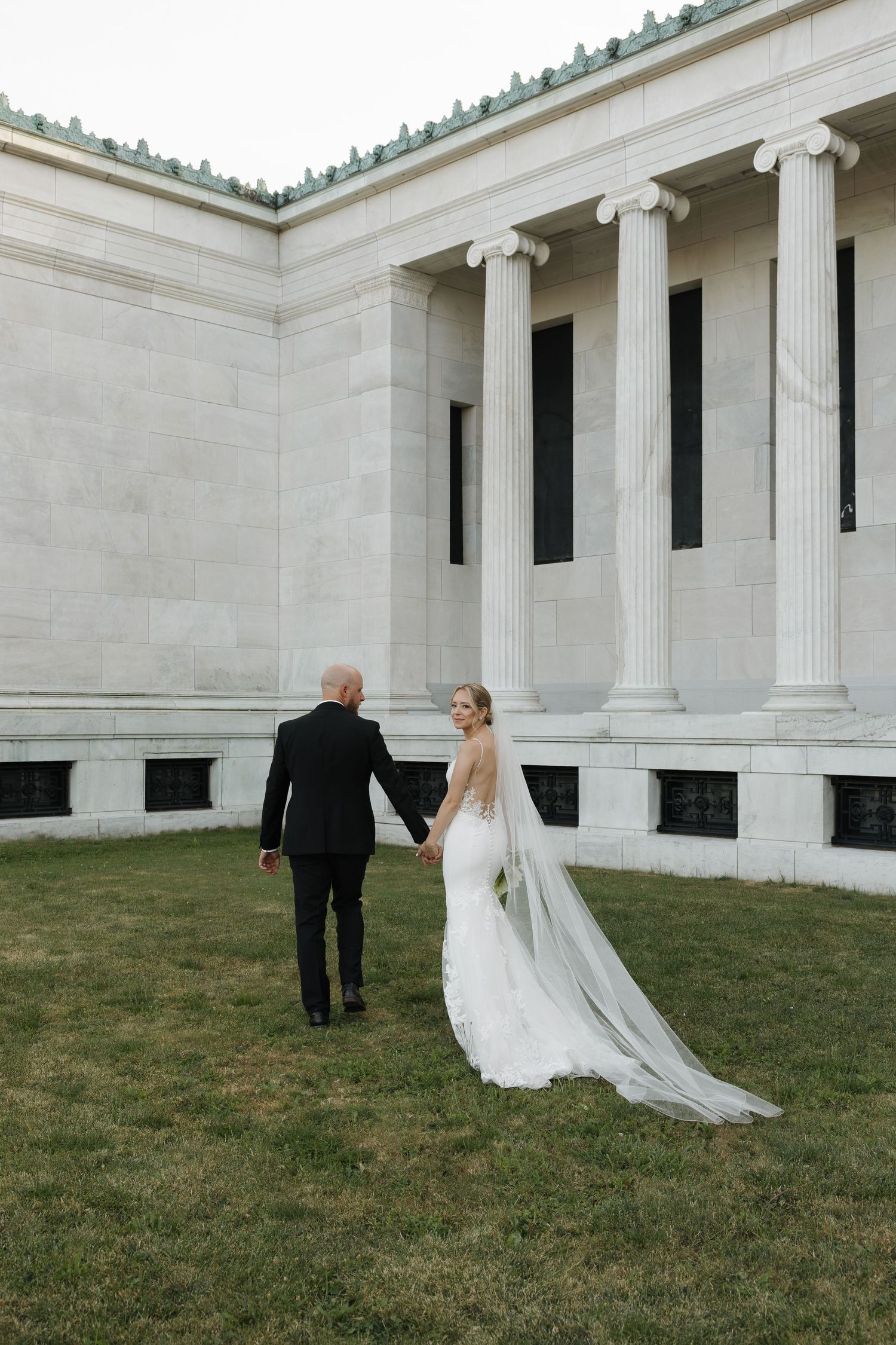 Bride and groom holding hands walking toward a grand neoclassical building with white columns on their wedding day.