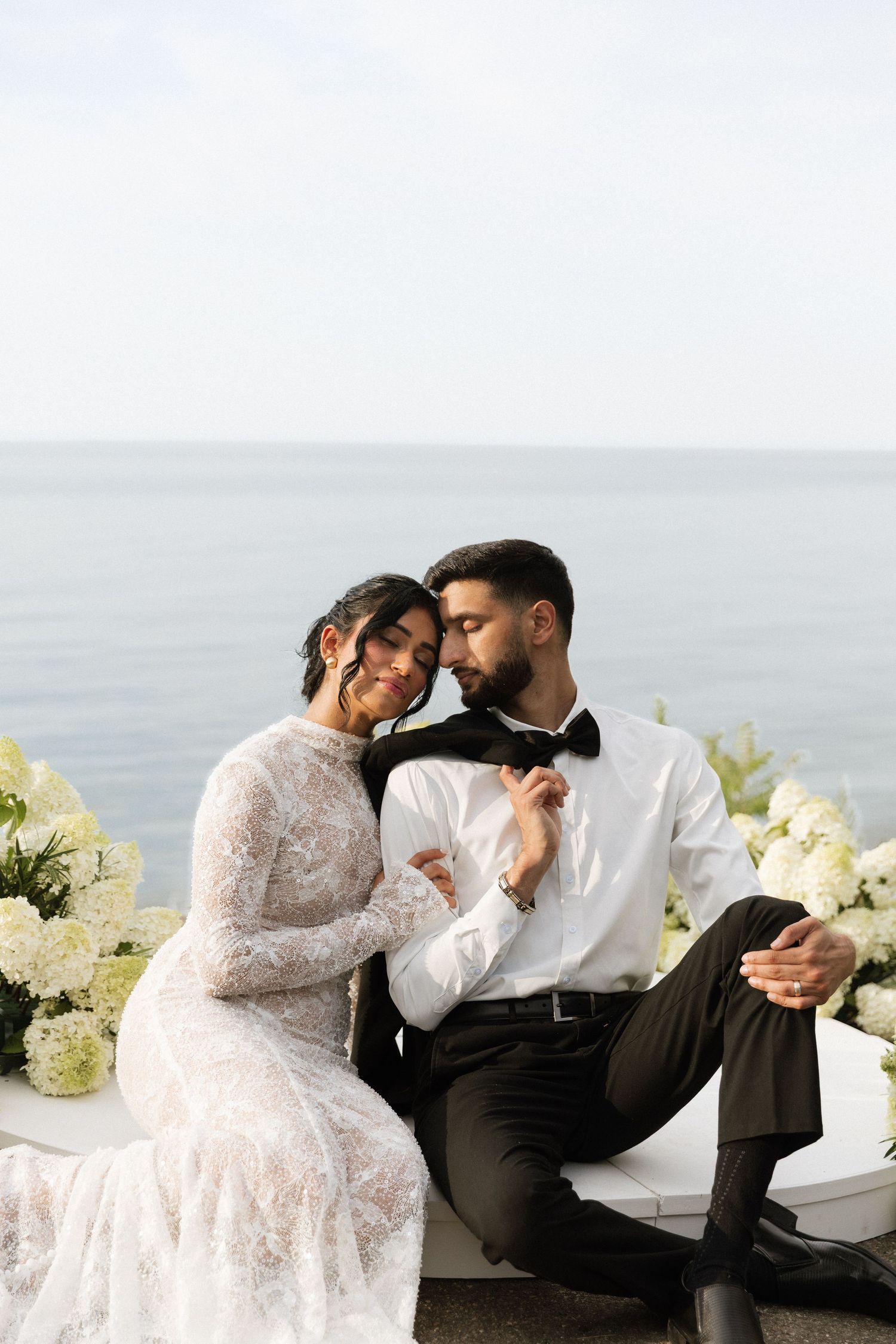 Romantic wedding couple embraces on seaside terrace with white floral decorations and ocean view backdrop.