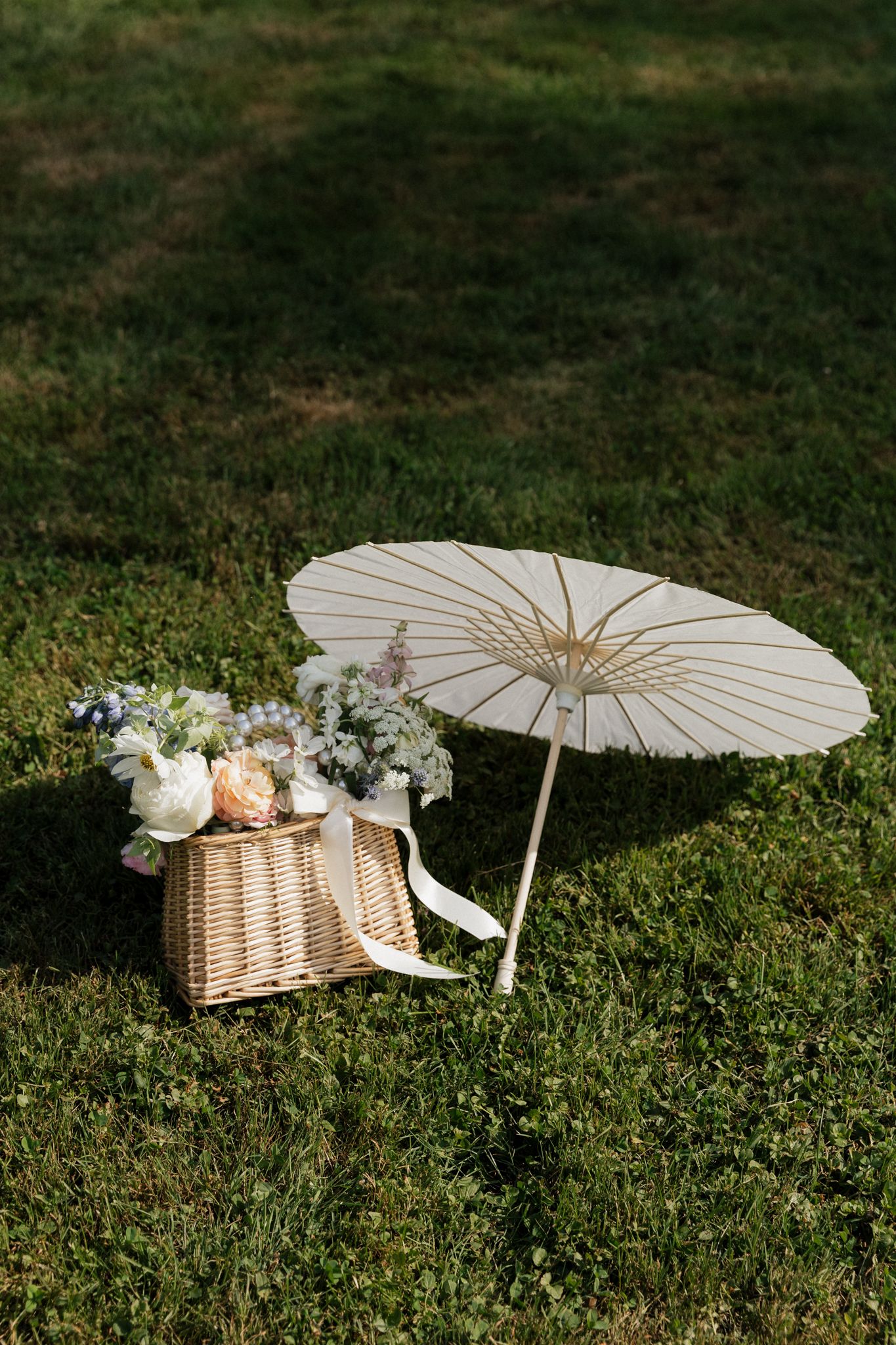 White paper parasol and wicker picnic basket with flowers sit on lush green grass in a romantic outdoor setting.