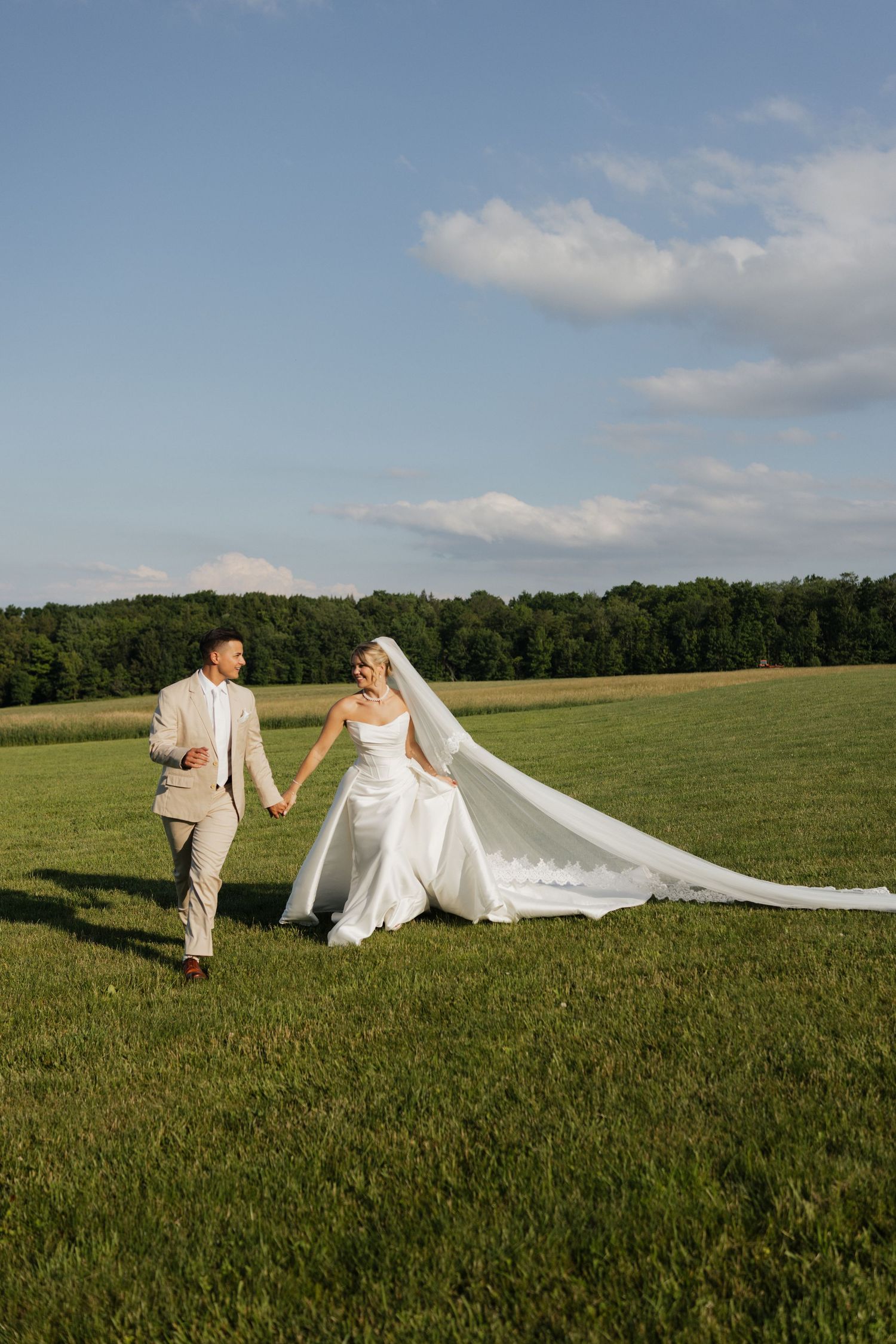 Bride and groom walk hand-in-hand across a lush green field, her veil flowing in the breeze under a blue sky.