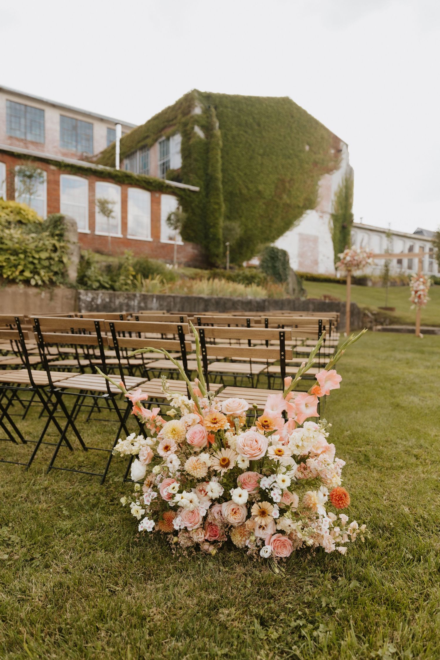 Outdoor wedding ceremony setup with pink and white floral arrangements lining wooden chairs on grass.