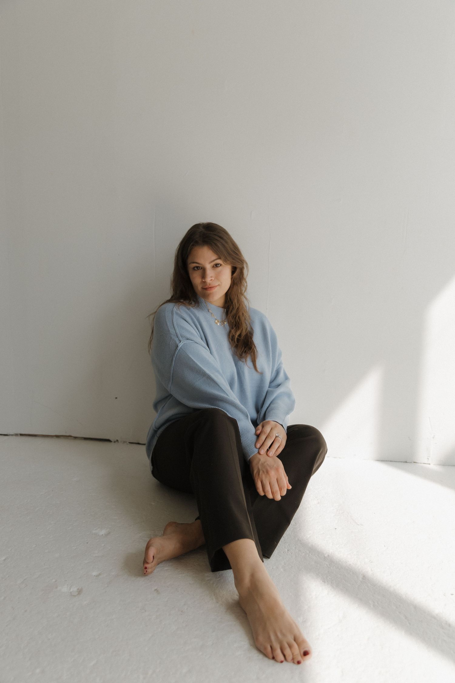 Person in light blue top and dark pants sits in minimalist white room with dramatic window lighting.