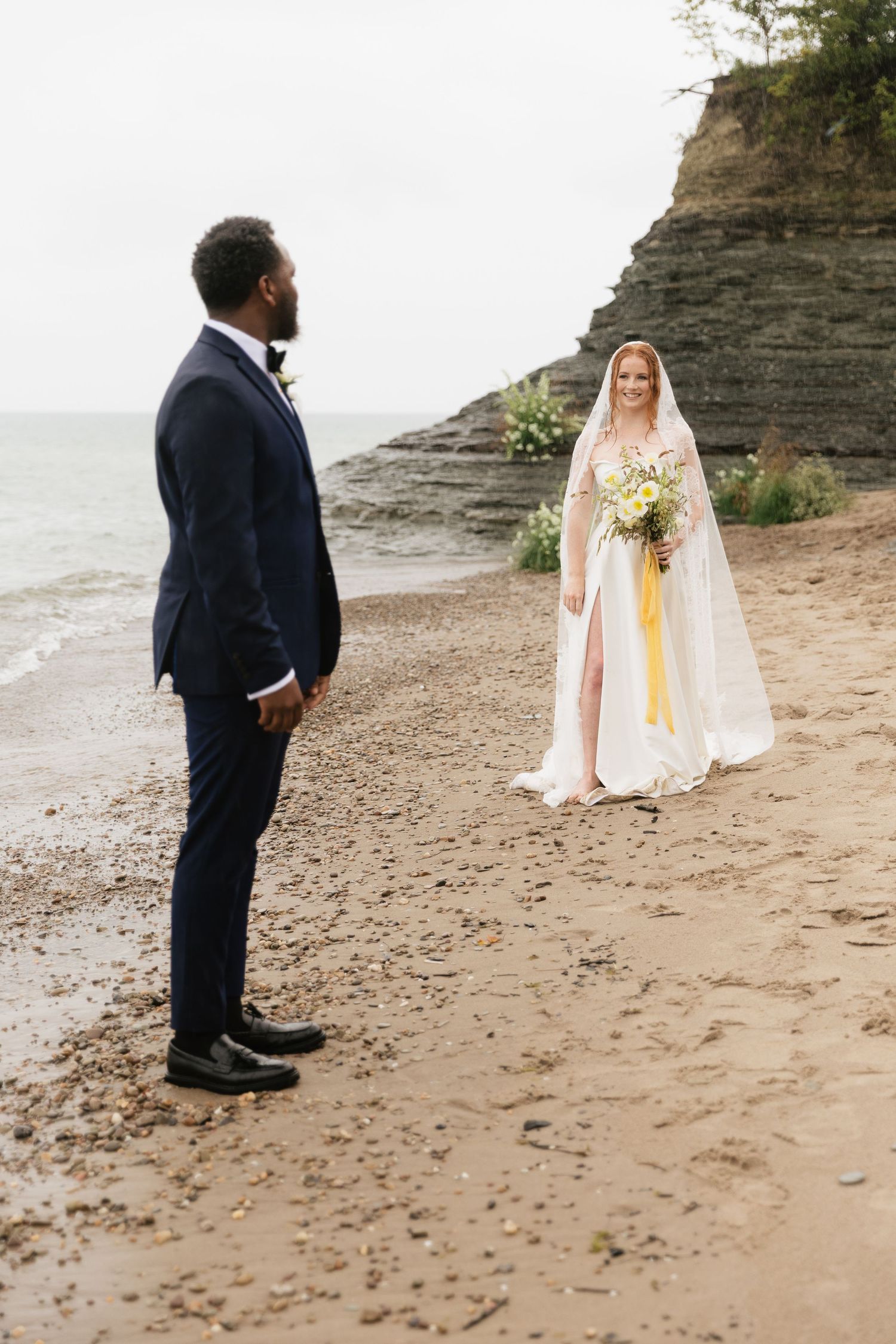 A couple shares a romantic moment on a sandy beach with dramatic cliffs in the background during their coastal wedding ceremony.