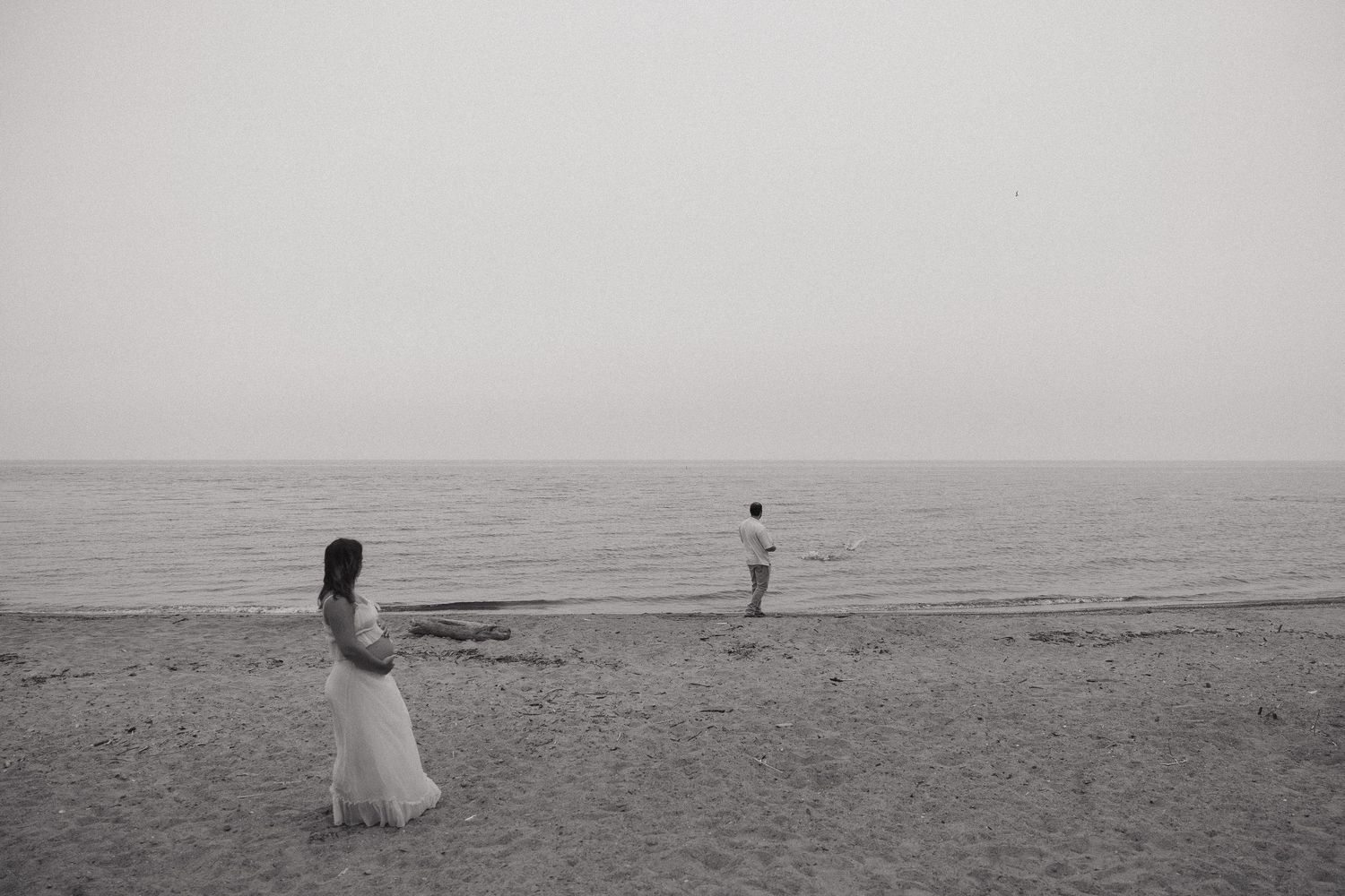 Black and white photo of a bride and groom on a beach near the ocean, standing apart in a romantic scene.