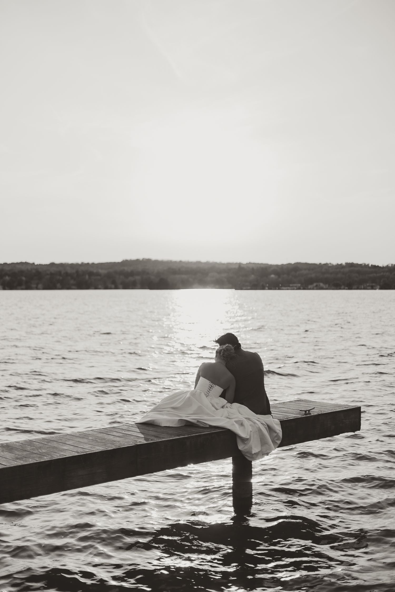 Black and white photo of people standing on a dock over water at sunset.