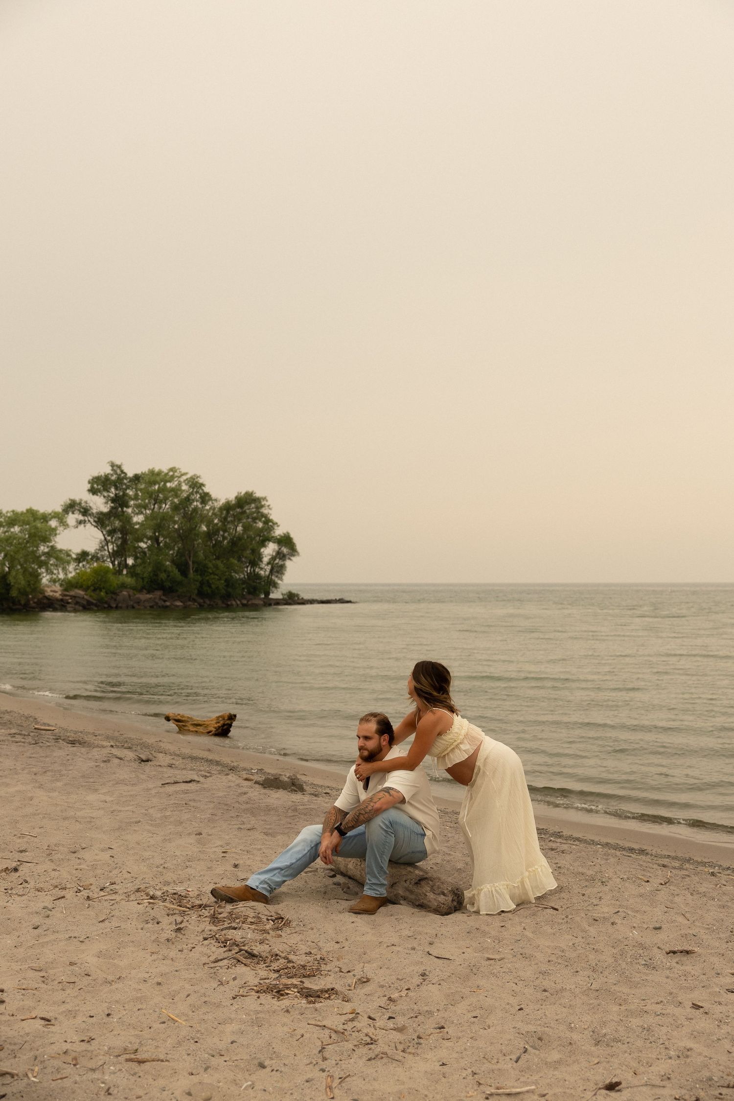 Couple in wedding attire sitting on a sandy beach shore with trees and calm water in the background.