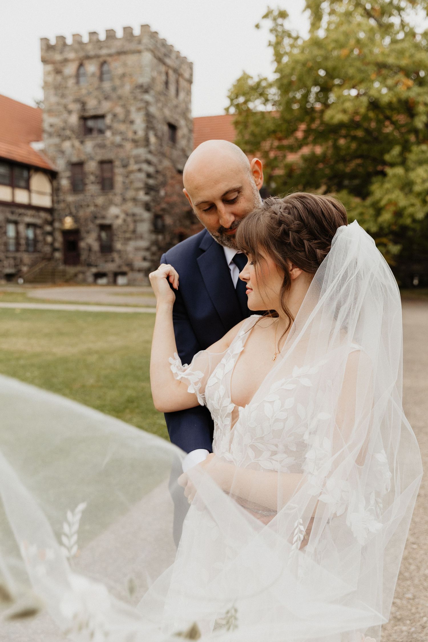 A romantic wedding couple embraces near a stone church building with a flowing white veil catching the autumn breeze.
