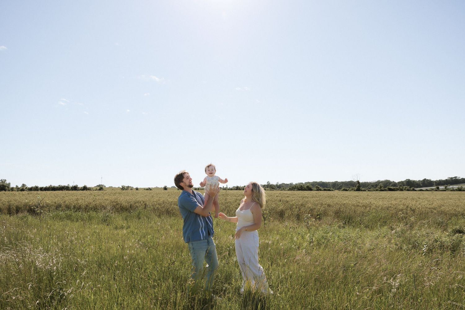 Family with baby enjoying sunny day in green field, parents lifting child up in the air outdoors.