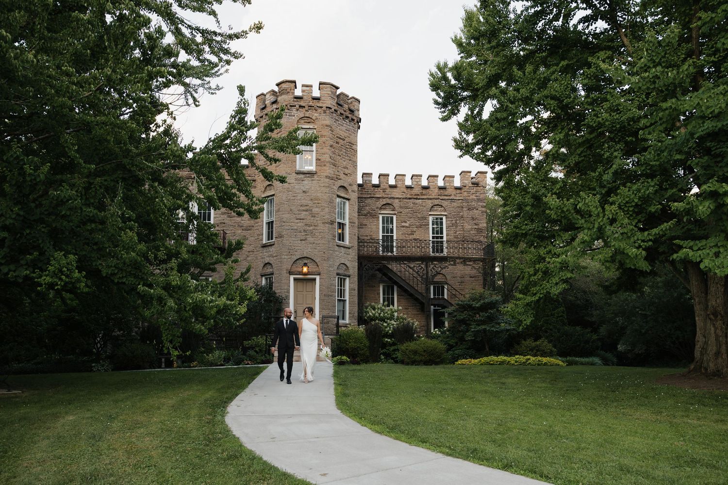 Bride and groom walk toward a historic stone castle venue surrounded by lush green trees on their wedding day.