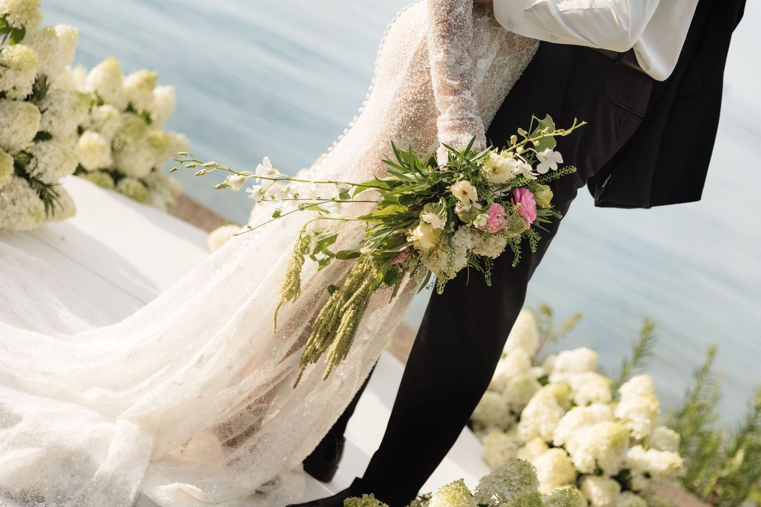A bride and groom share a romantic embrace during their outdoor wedding ceremony with white floral decorations.