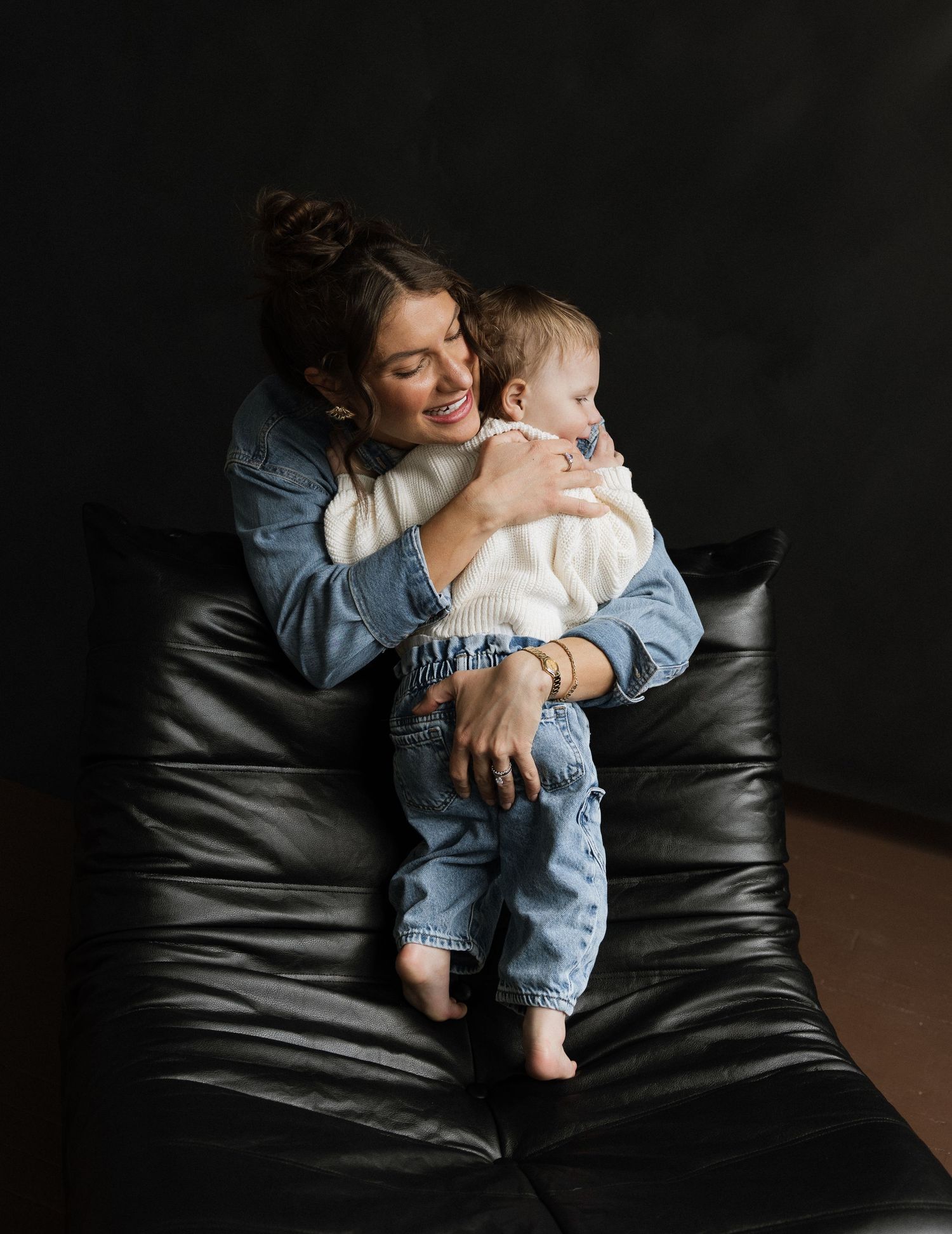 A smiling woman in a denim jacket holds a happy baby on a black leather chair against a dark background.