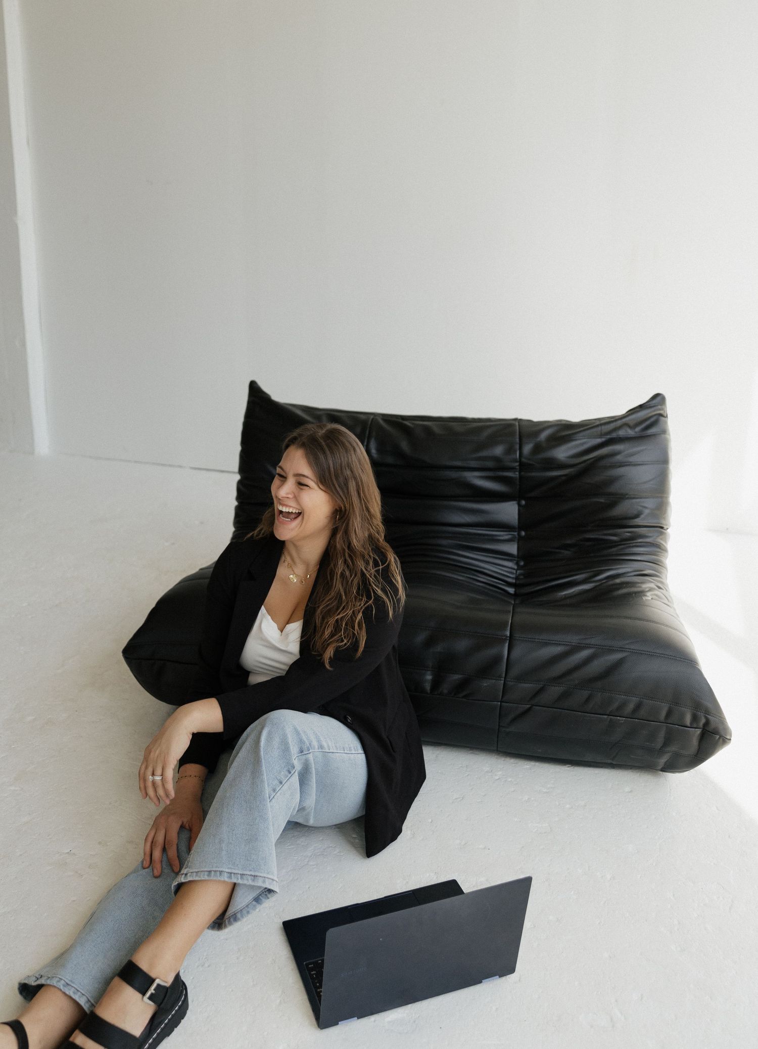 Woman in casual attire lounges on black leather cushions while laughing and relaxing in a bright white room.