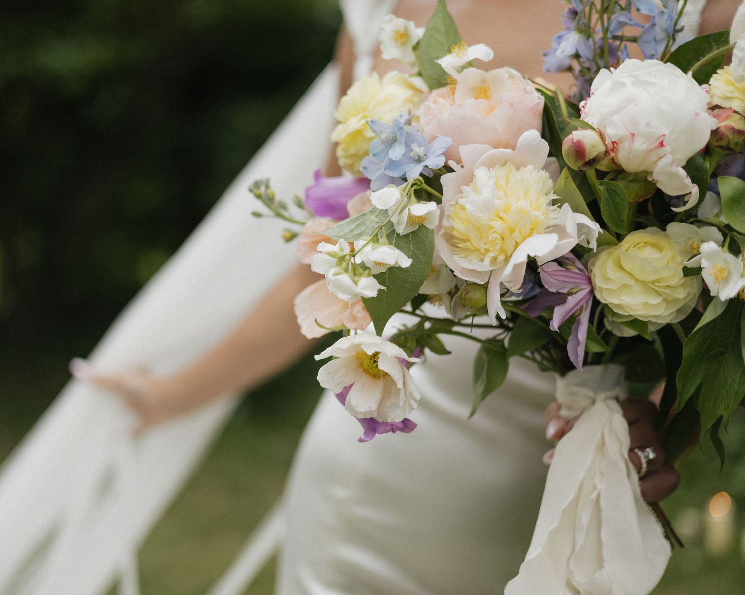 A romantic wedding bouquet featuring soft pink and cream roses with delicate purple flowers against a white dress.
