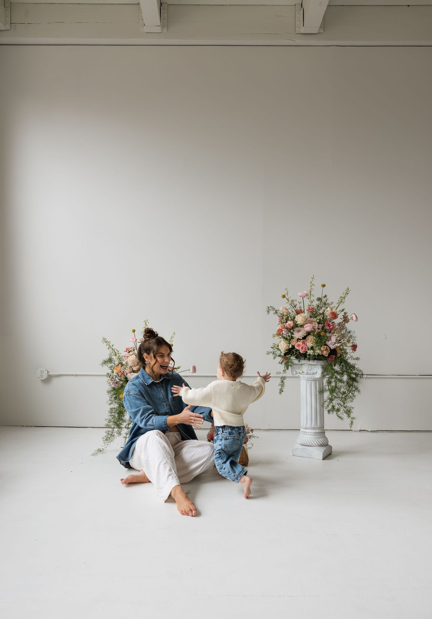 Adult sitting on white floor with toddler near floral arrangements on pedestal in bright minimalist studio.