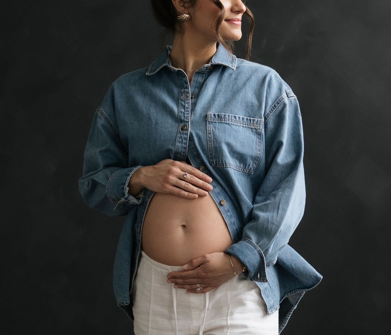 Pregnant person wearing unbuttoned denim shirt shows bare belly against dark background in artistic maternity photo.