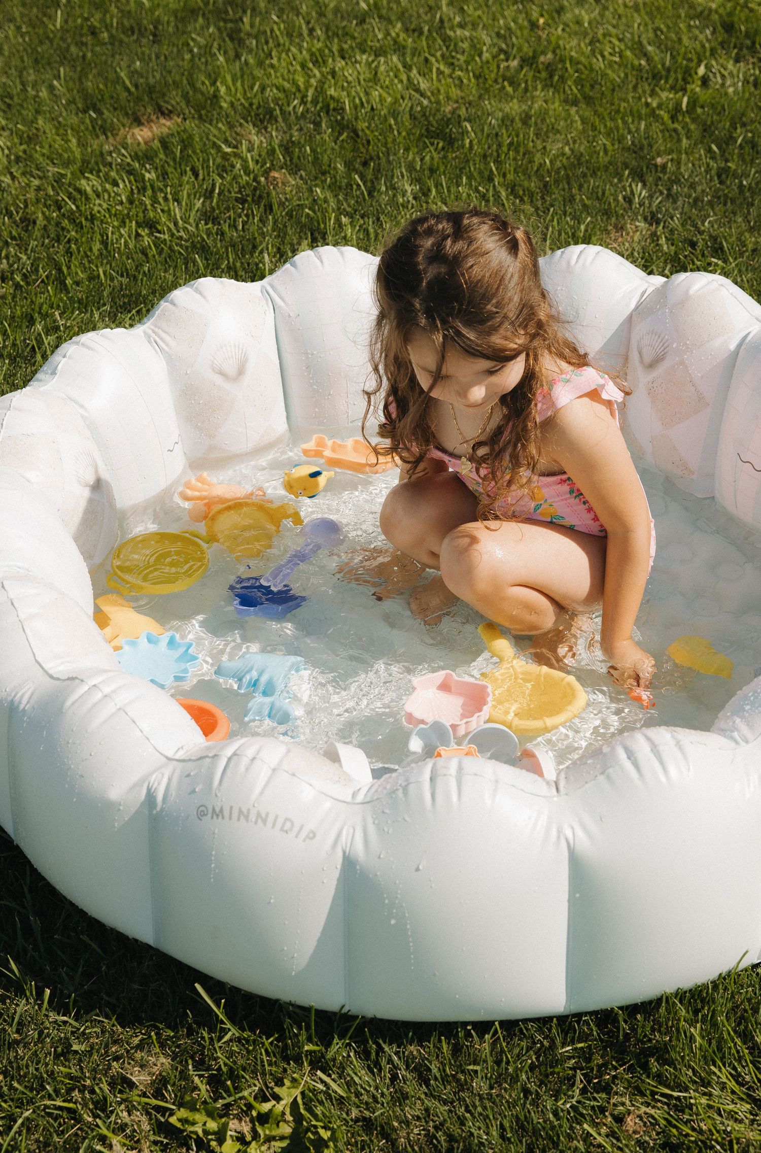 Young girl playing in a Minnidip inflatable kiddie pool on green grass, surrounded by colorful water toys.