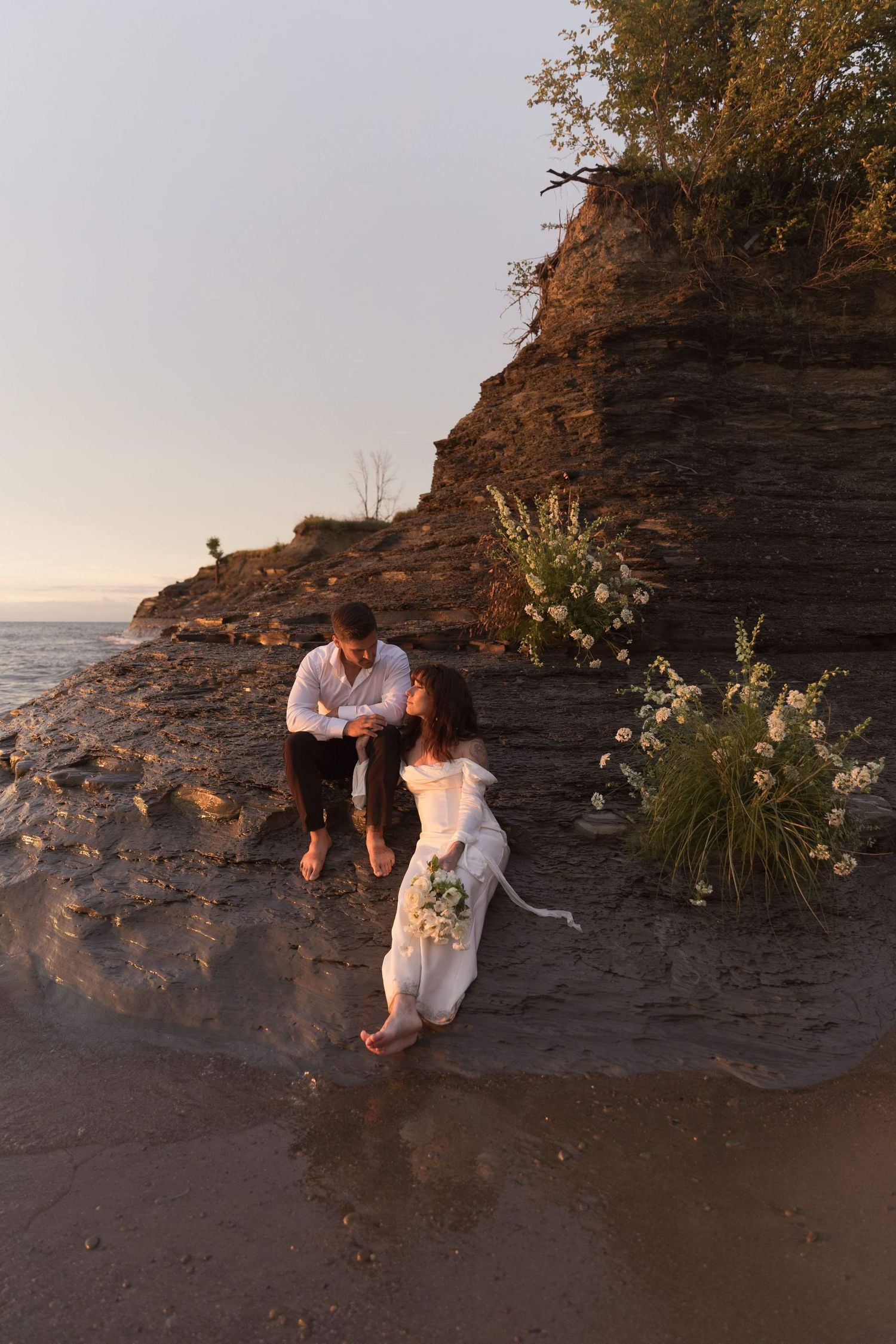 A couple shares a romantic moment on a rocky beach at sunset with floral decorations against a dramatic cliff backdrop.