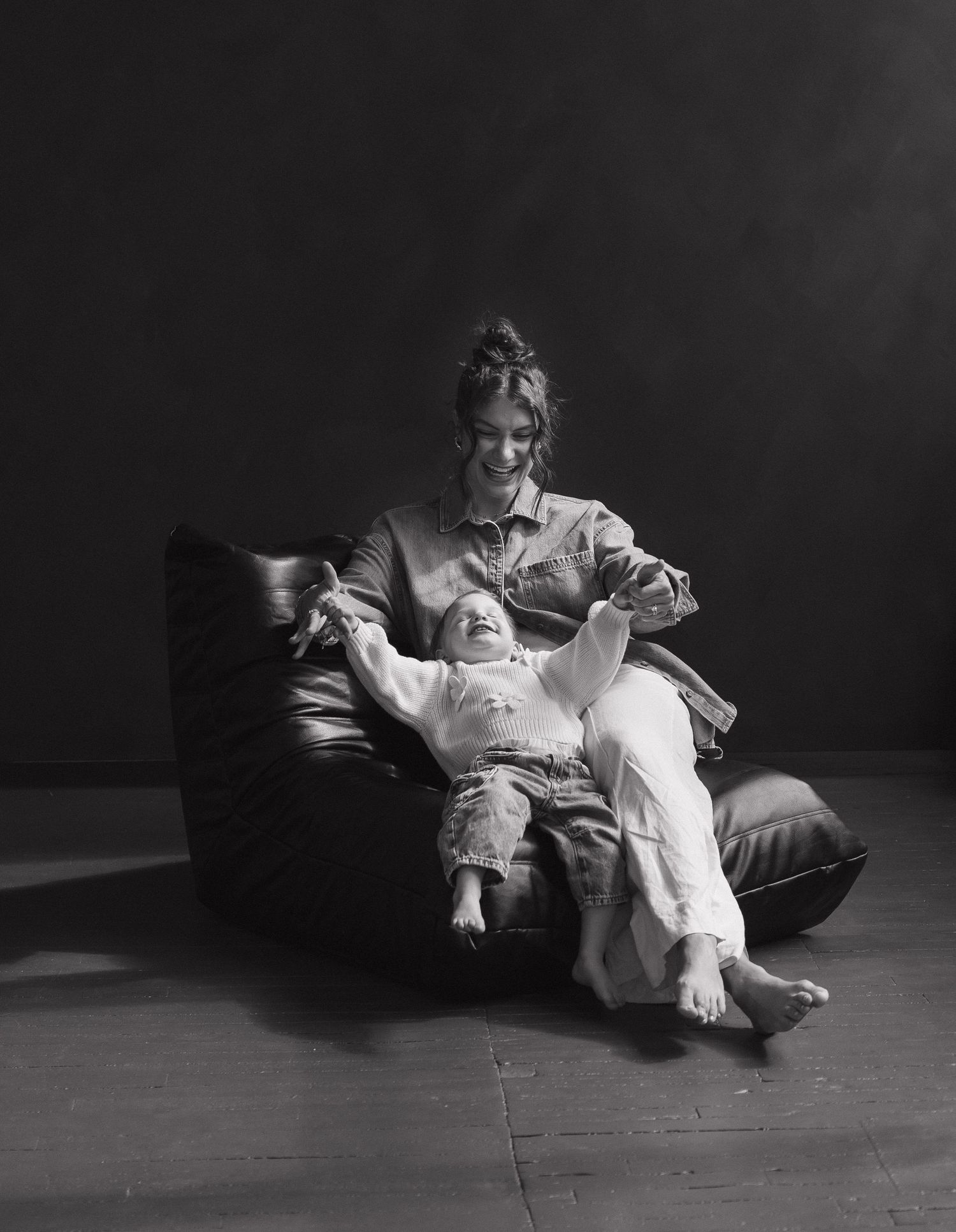 Black and white photo of a smiling woman and laughing child relaxing together on a leather bean bag chair.