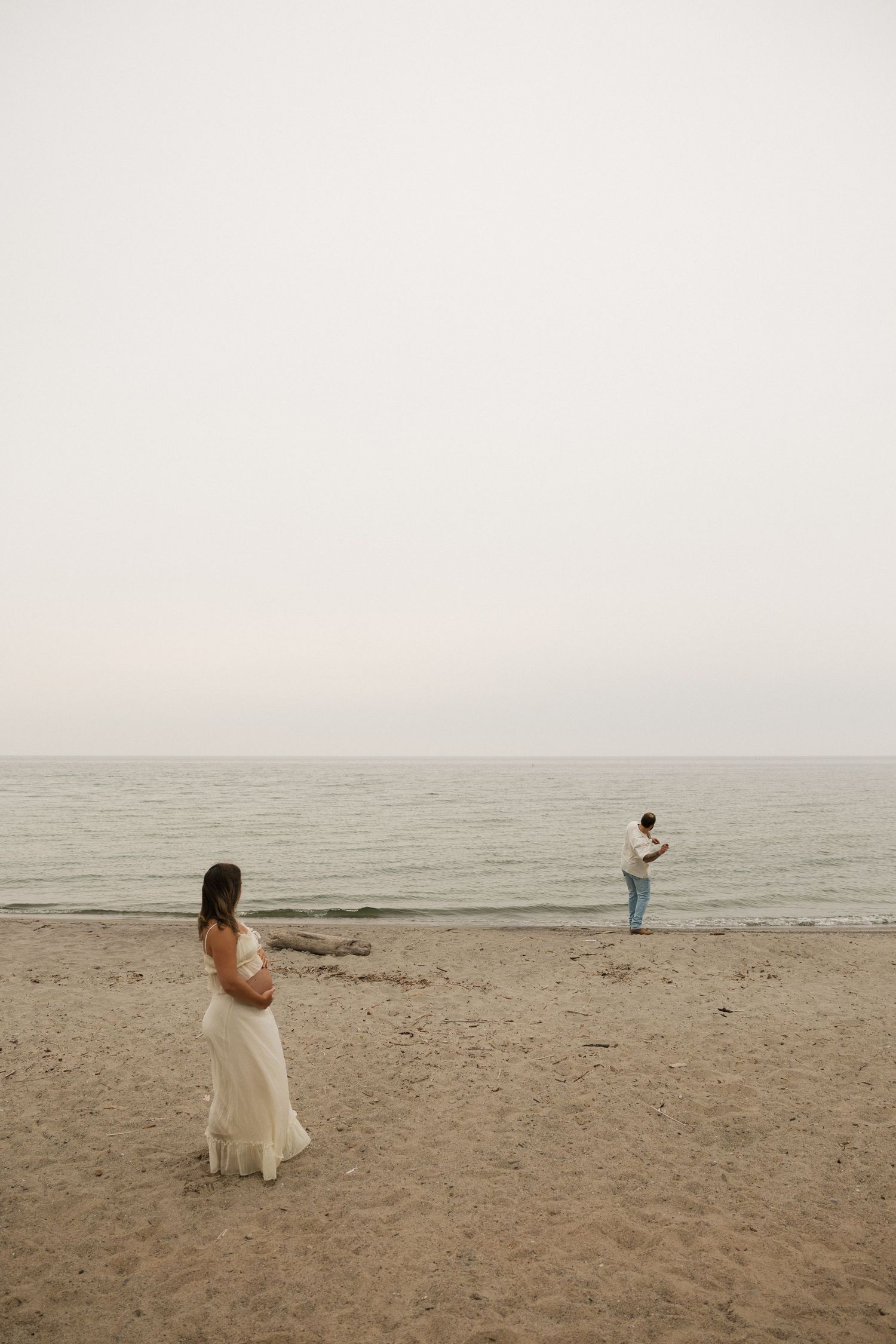 Pregnant woman in white dress stands on sandy beach while partner gazes at calm ocean on overcast day.