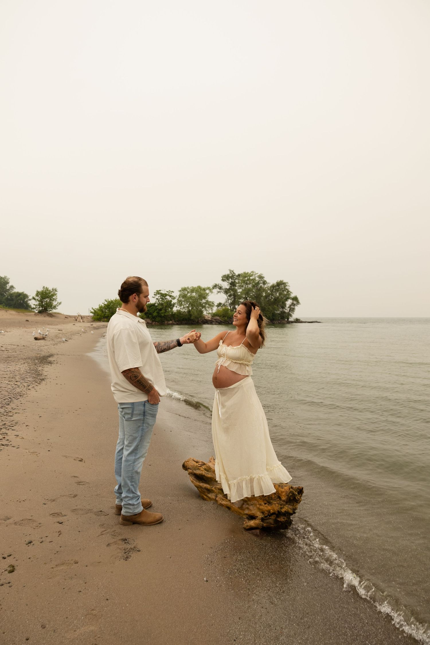 Pregnant couple holding hands on a sandy beach shoreline, woman in white dress standing on rock, man in jeans and white shirt.
