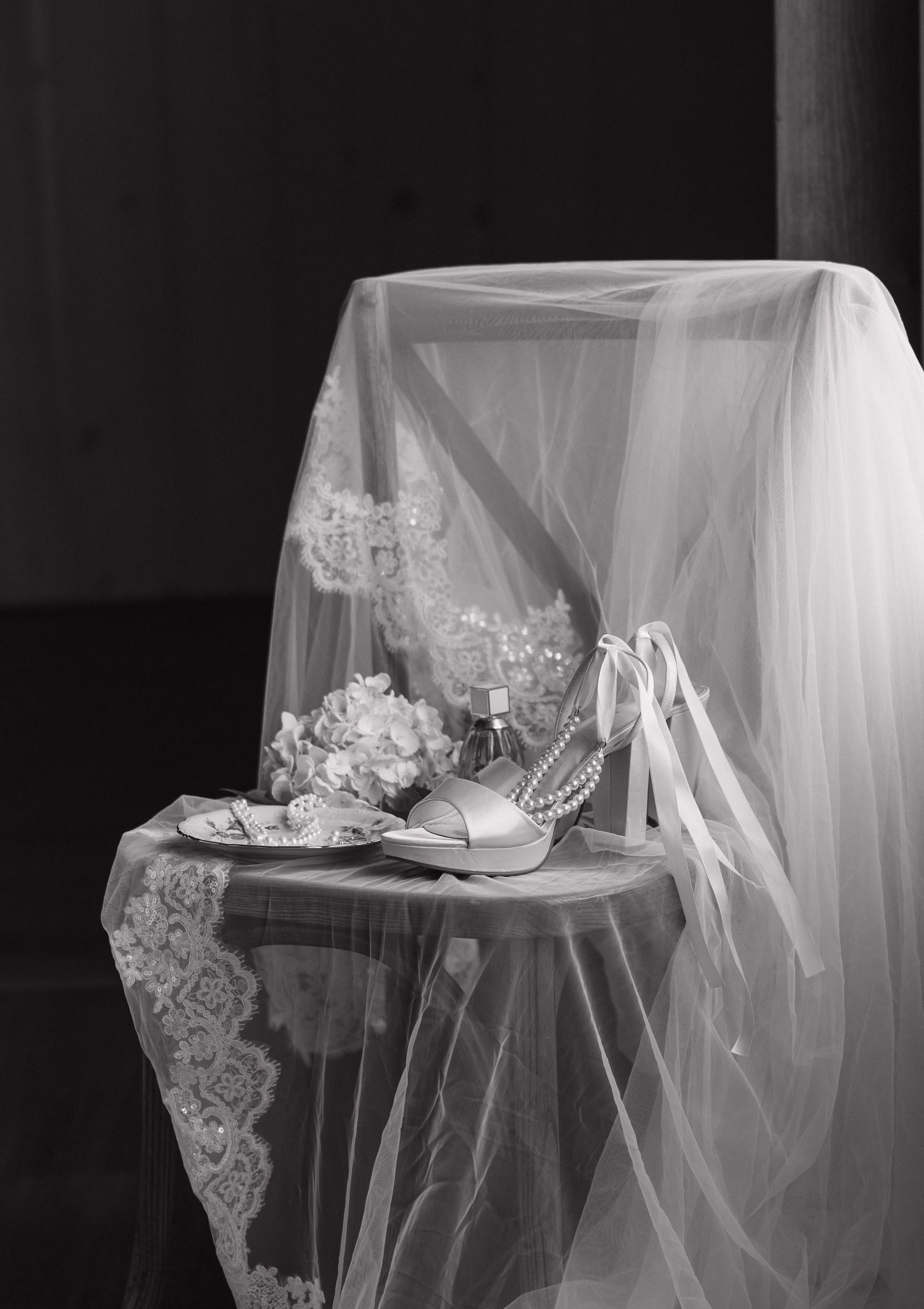 Black and white photo of bridal accessories including heels, pearls, bouquet, and lace veil draped over a small table.