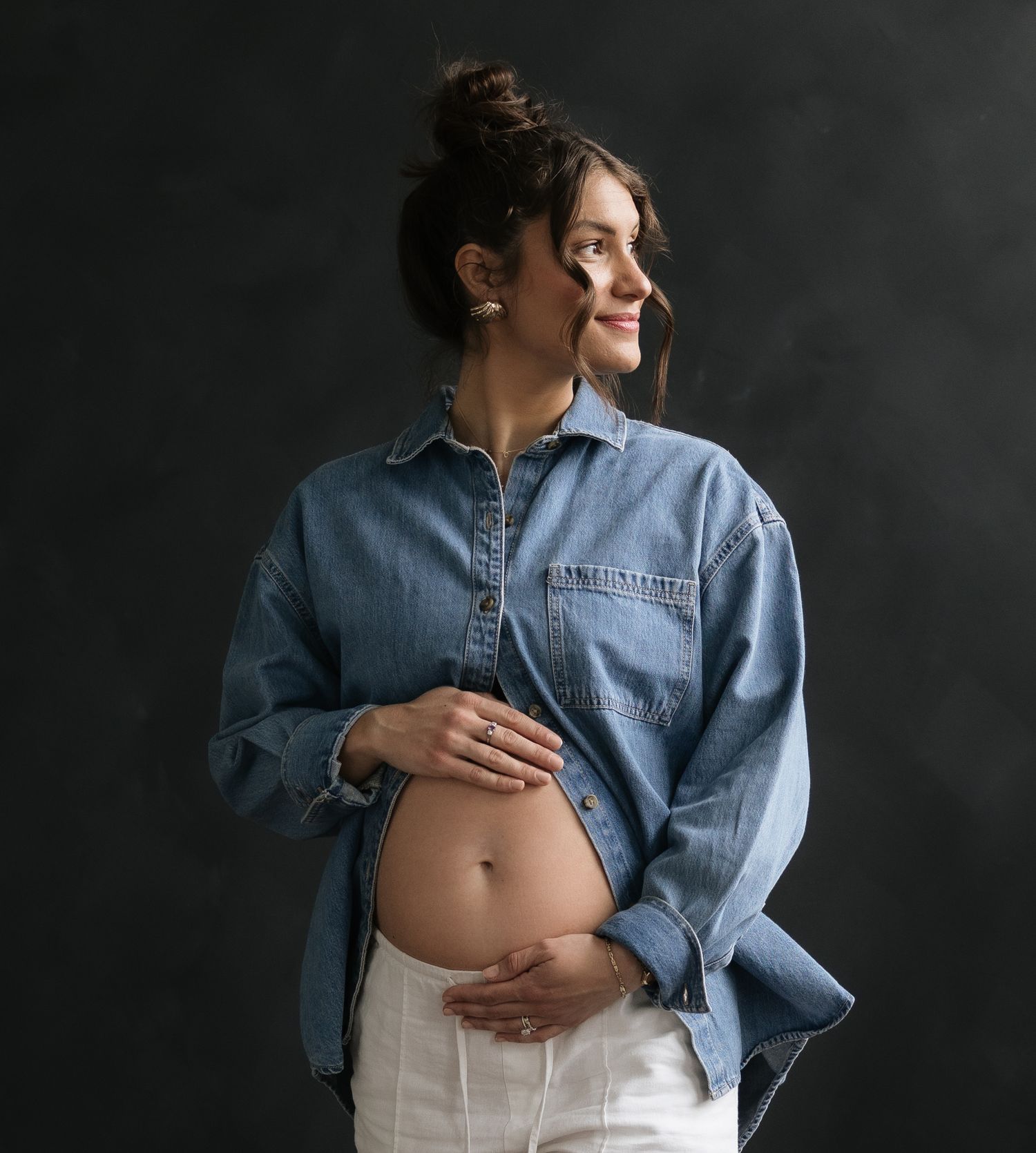 Smiling pregnant woman in denim shirt cradling baby bump against dark background.