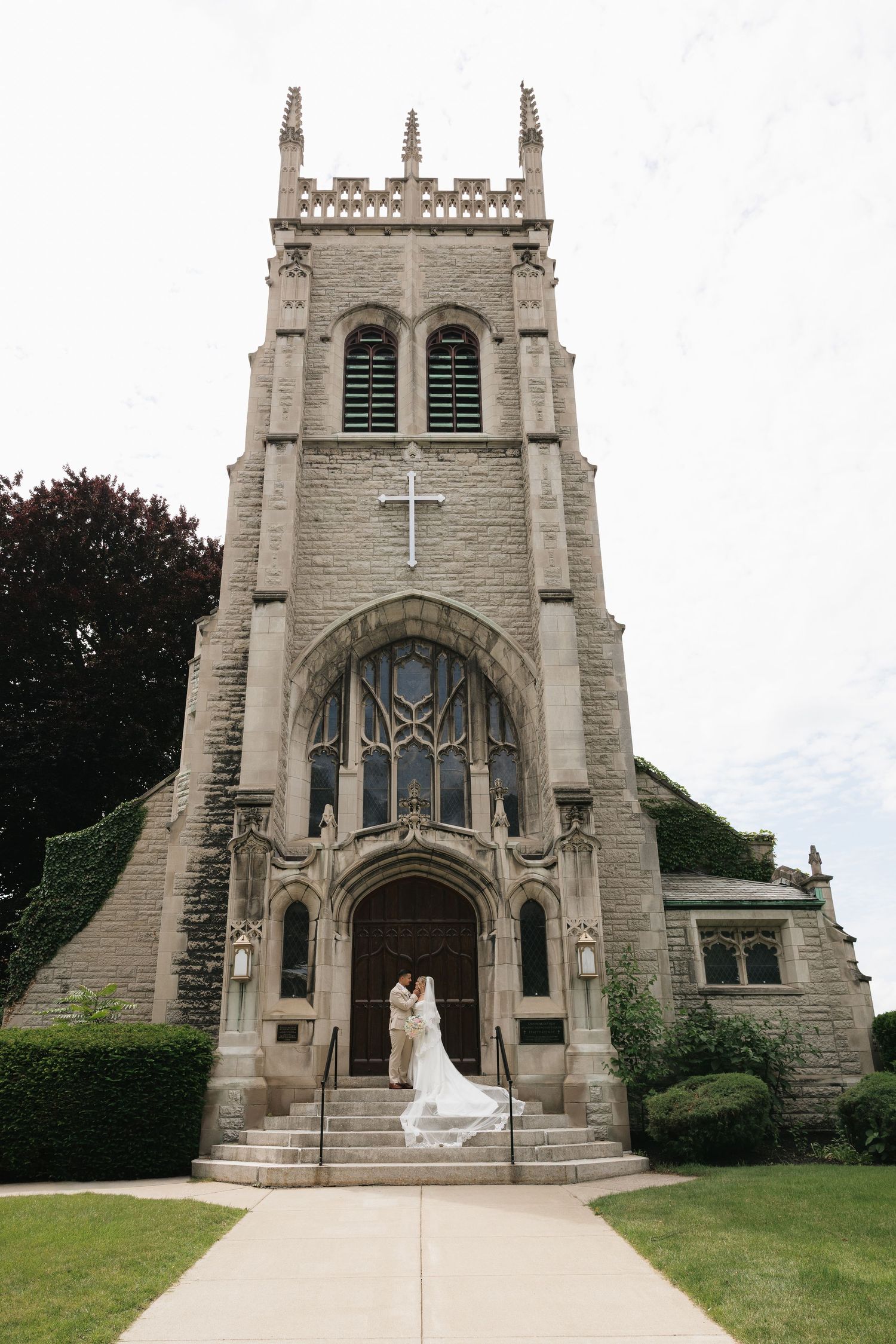 Bride and groom embrace at the entrance of a stunning Gothic stone church with a cross and tower on their wedding day.