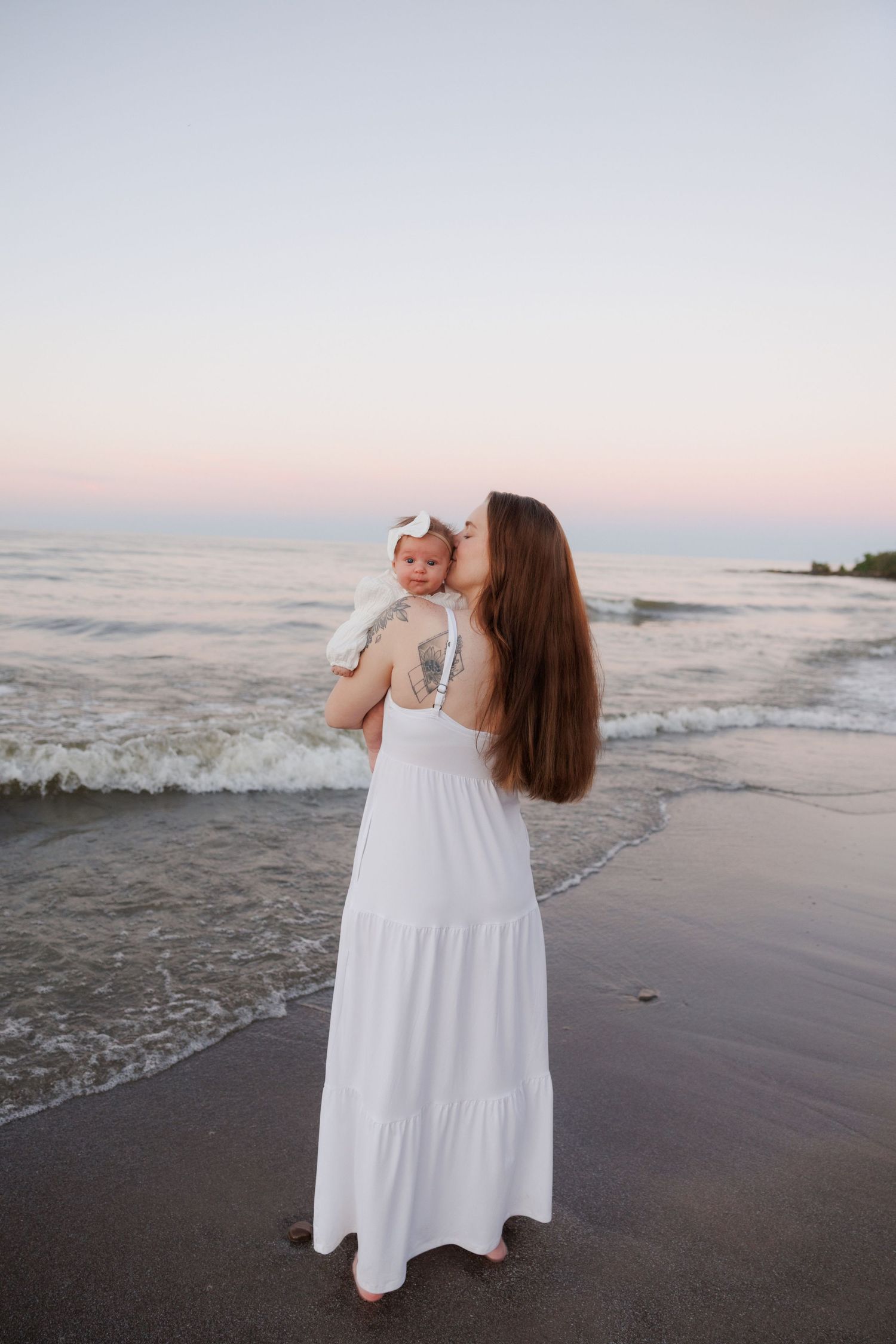 Mother in white maxi dress kissing baby girl on beach at sunset, standing at beach edge.