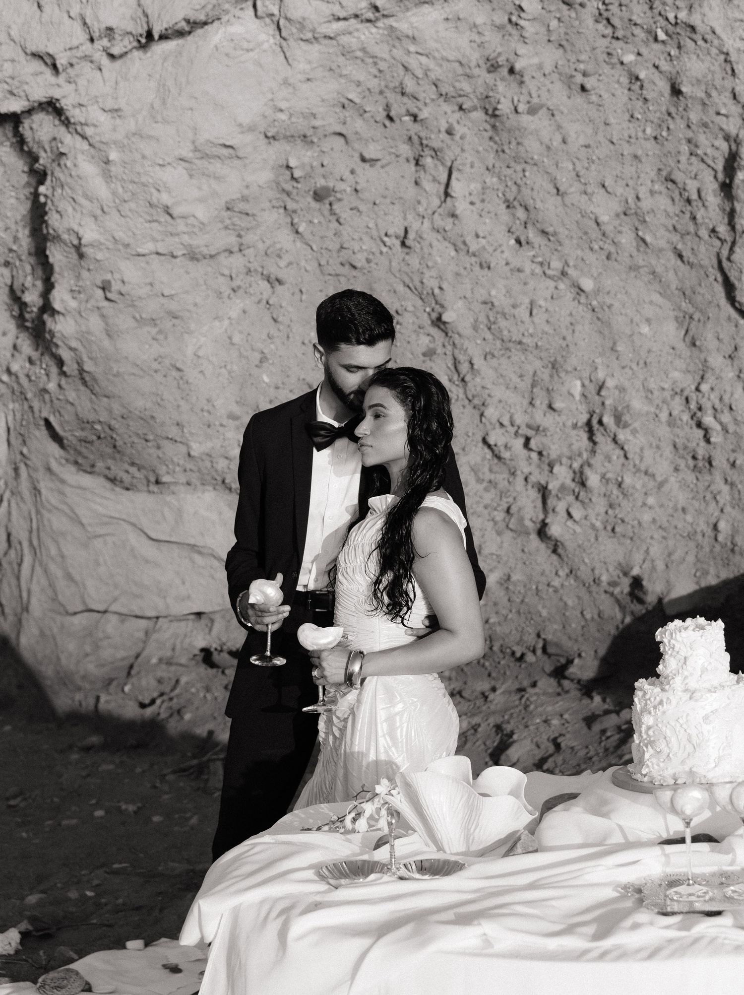Black and white wedding photo series showing a couple sharing cake and champagne against a textured stone wall backdrop.