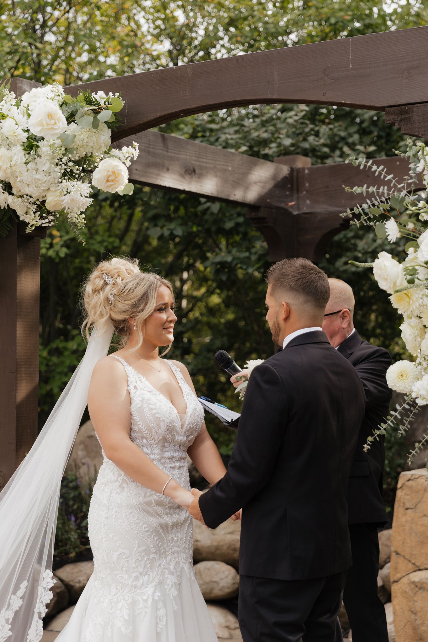 Wedding ceremony taking place under a wooden pergola decorated with white floral arrangements and greenery.