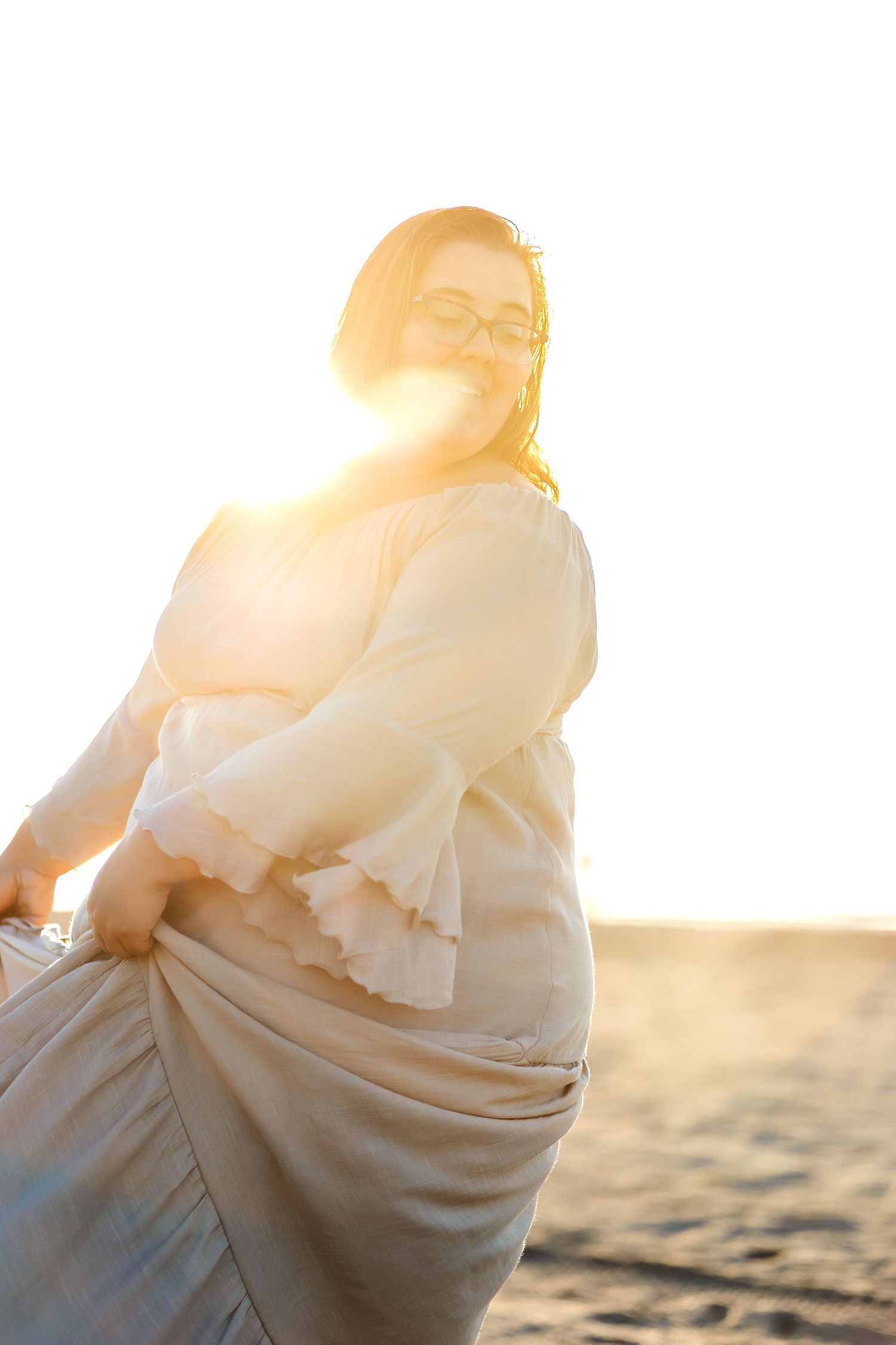 Thalie photography Woman in flowing white dress holding skirt at golden hour sunset on sandy beach with bright backlight.
