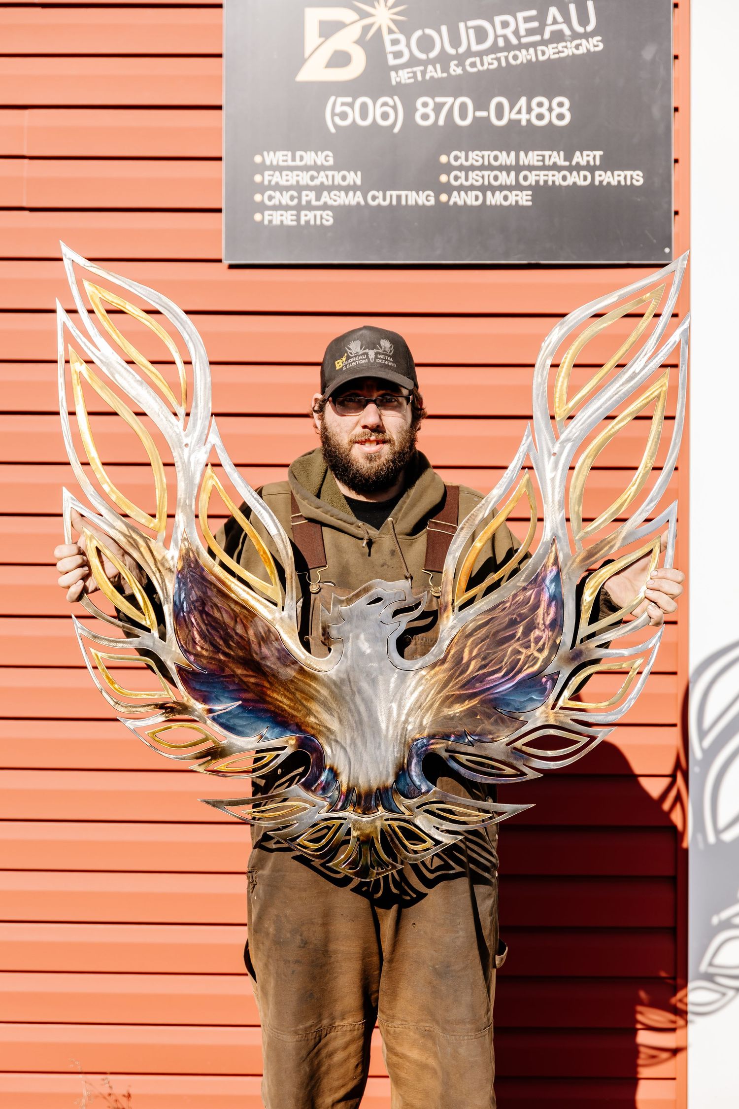 A bearded man holds a large colorful metal phoenix art piece outside Boudreau Metal & Custom Designs shop.