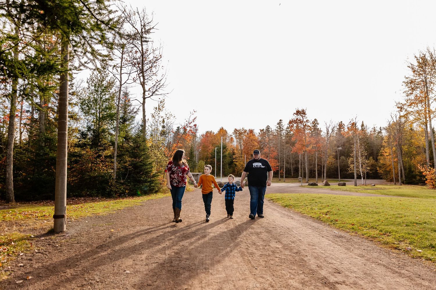 Family of four walking hand-in-hand down a scenic dirt path surrounded by colorful autumn trees.