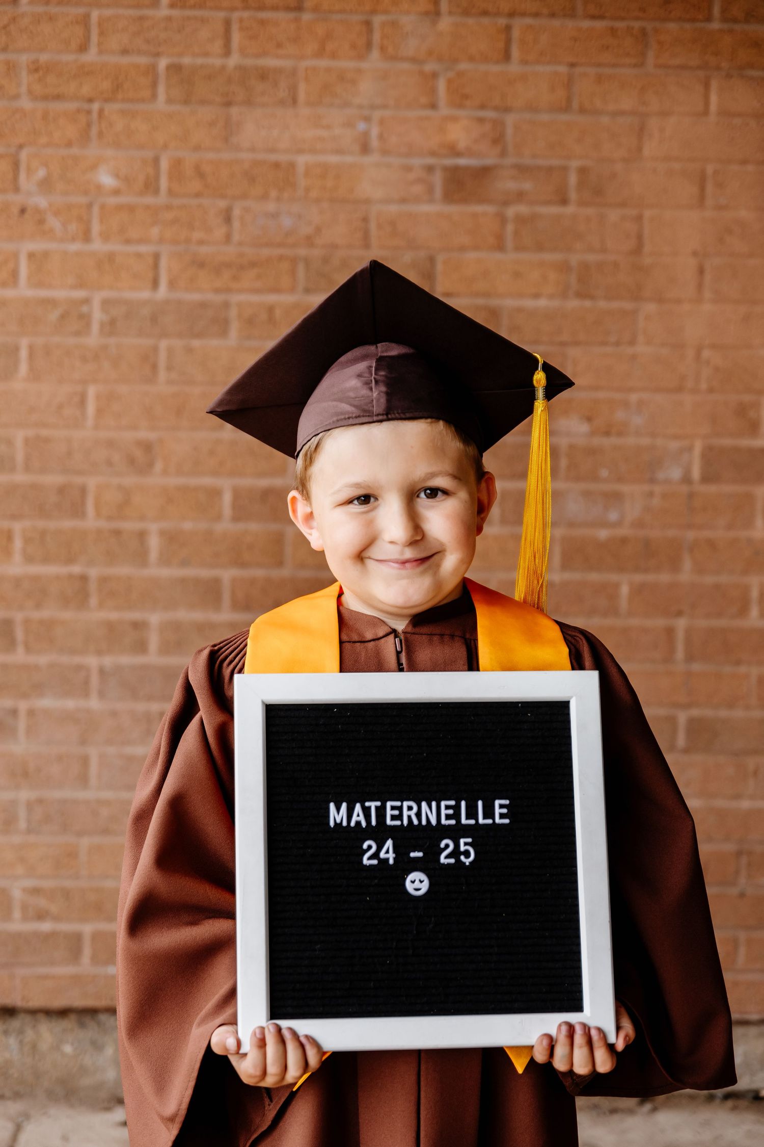 Happy young boy in brown graduation cap and gown holding a letterboard reading Maternelle 24-25.