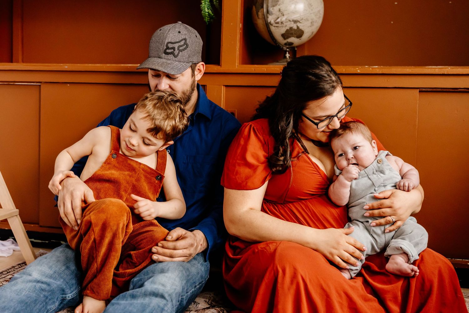 Family portrait of parents with toddler son and newborn baby seated against a warm orange wall.