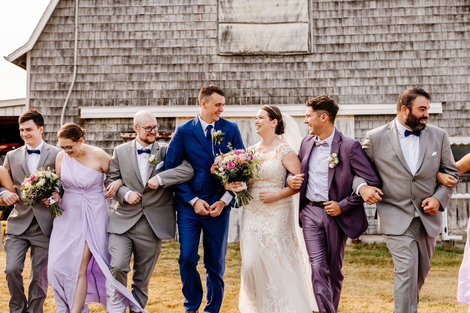 Wedding party walking and laughing together in front of a rustic barn in coordinated gray and purple attire.