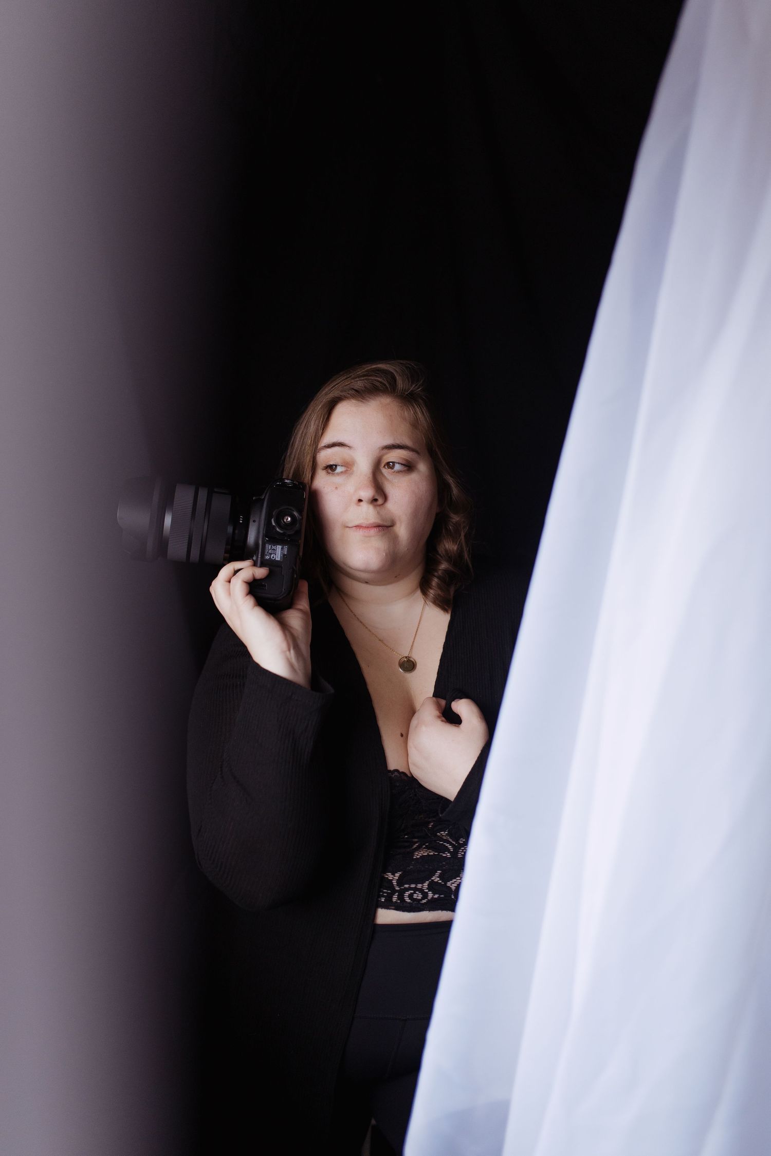 Thalie photography Female photographer holding DSLR camera peeking through dark studio backdrop beside white curtain.