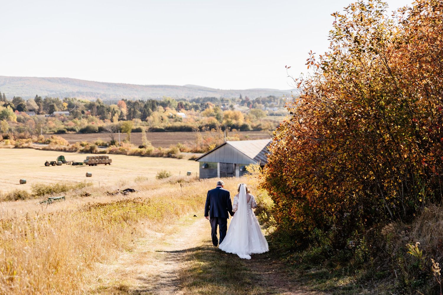 Bride and groom walking hand in hand along a rural path in autumn countryside.