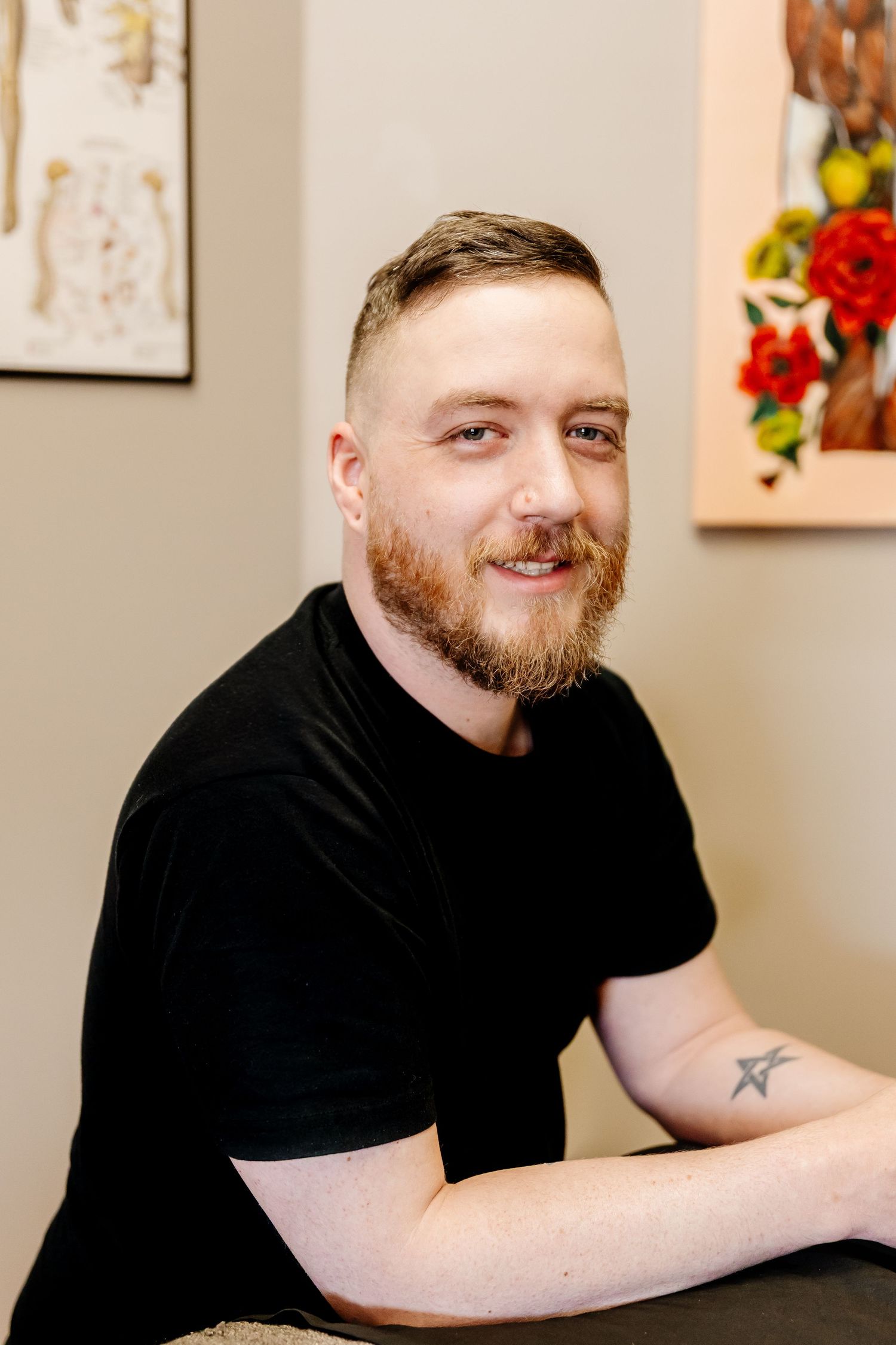 Smiling man with red beard and star tattoo on forearm wearing black t-shirt seated in a bright studio space.
