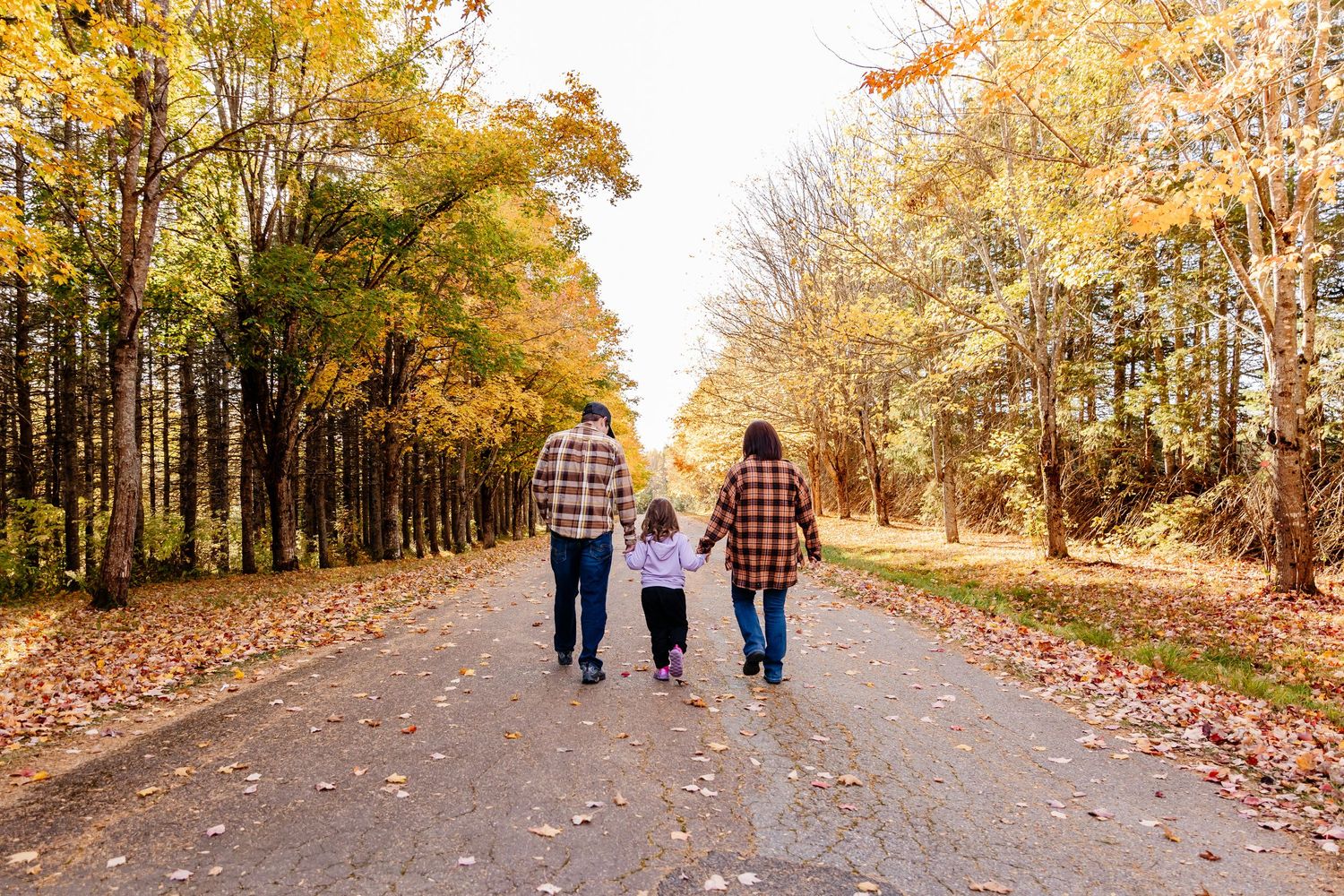 Family of three walking hand-in-hand down a scenic autumn tree-lined path covered with fallen leaves.